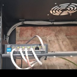 BPZ - Switch sieciowy BCS zamontowany w szafie rack, z podłączonymi kablami Ethernet, widoczne diody sygnalizujące pracę portów PoE, kable ułożone w korytkach, ściana z cegły w tle.