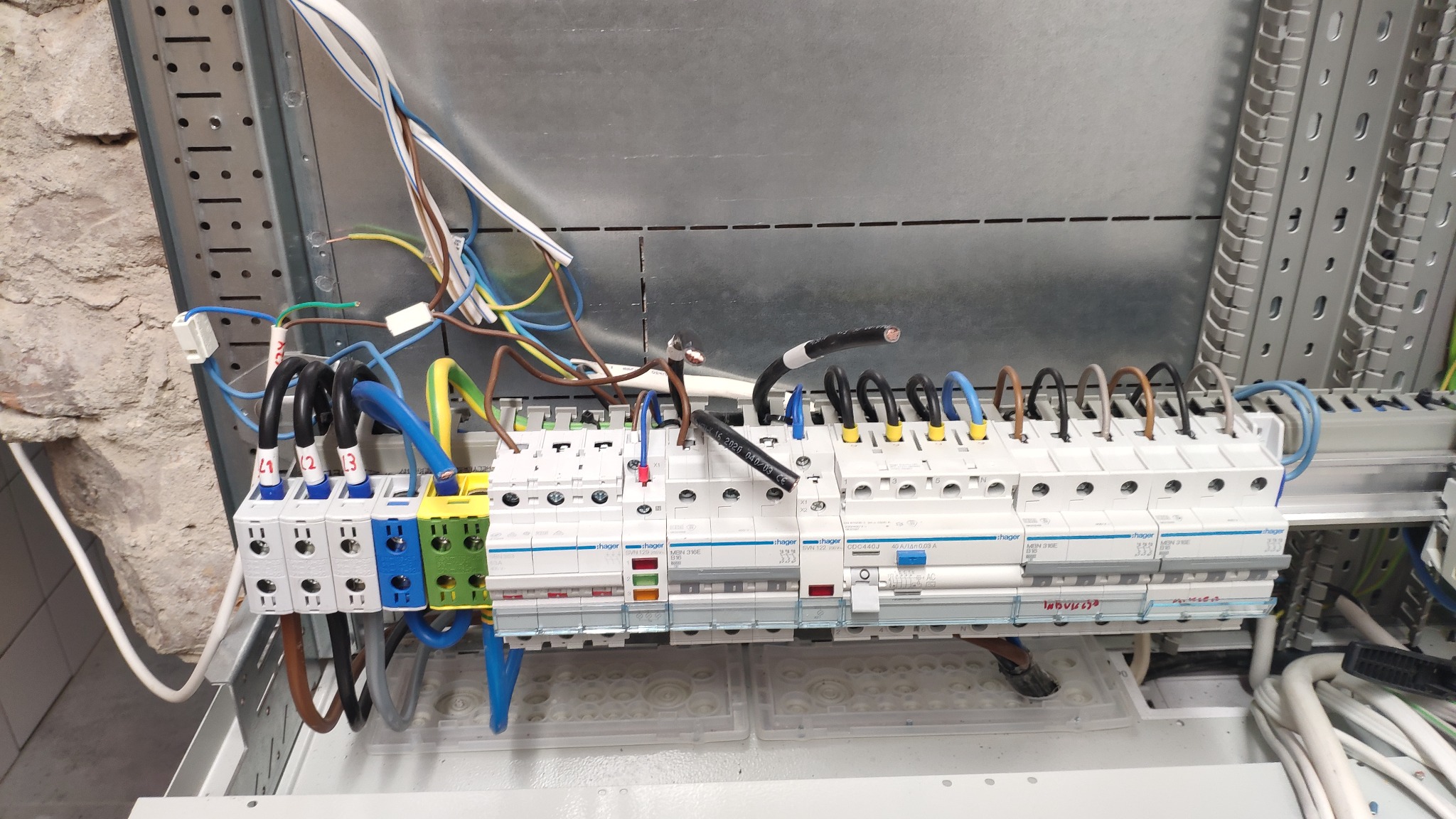 Rozdzielnica elektryczna w trakcie montażu, widoczne przewody z oznaczeniami L1, L2, L3, bezpieczniki Hager, okablowanie w kolorach brązowym, niebieskim, czarnym i żółto-zielonym, instalacja...