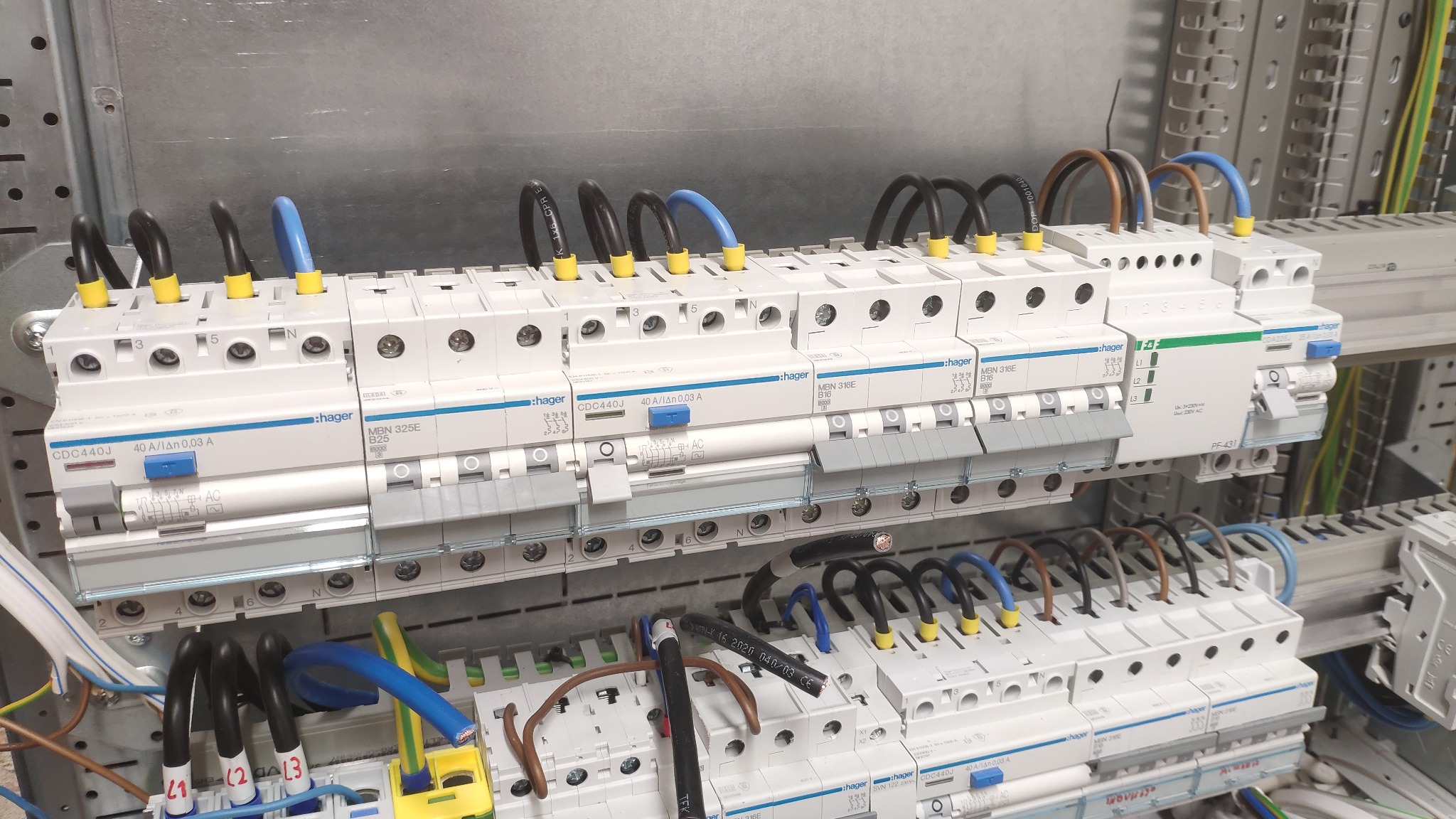 Szyna DIN z zamontowanymi bezpiecznikami różnicowoprądowymi Hager, widoczne przewody w kolorach czarnym, niebieskim i brązowym, z żółtymi oznaczeniami na końcówkach, ułożone w górnej i dolnej...