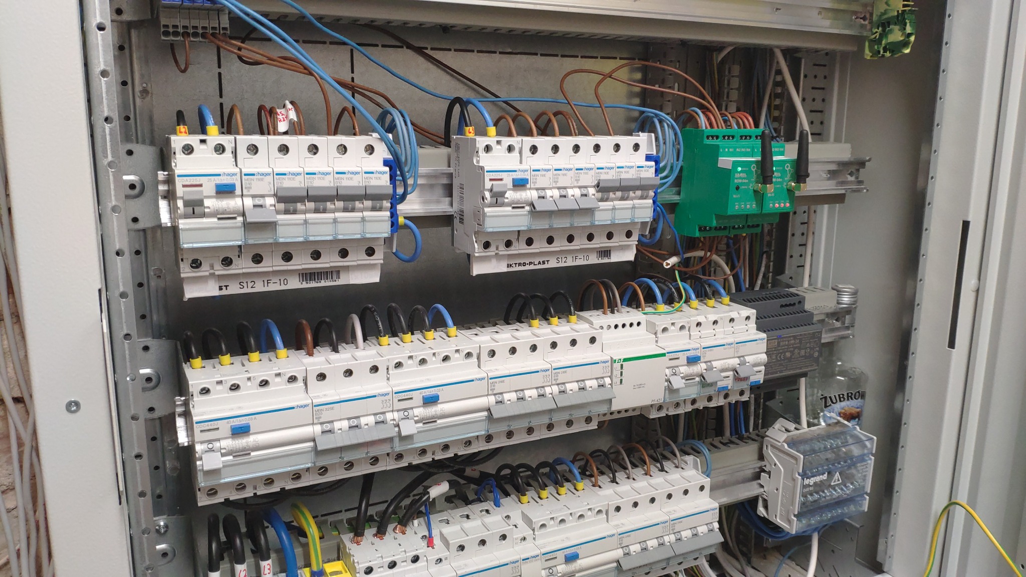 Rozdzielnica elektryczna z zamontowanymi bezpiecznikami Hager, przewodami w różnych kolorach i modułem sterującym, widoczna butelka Żubrówki w tle.