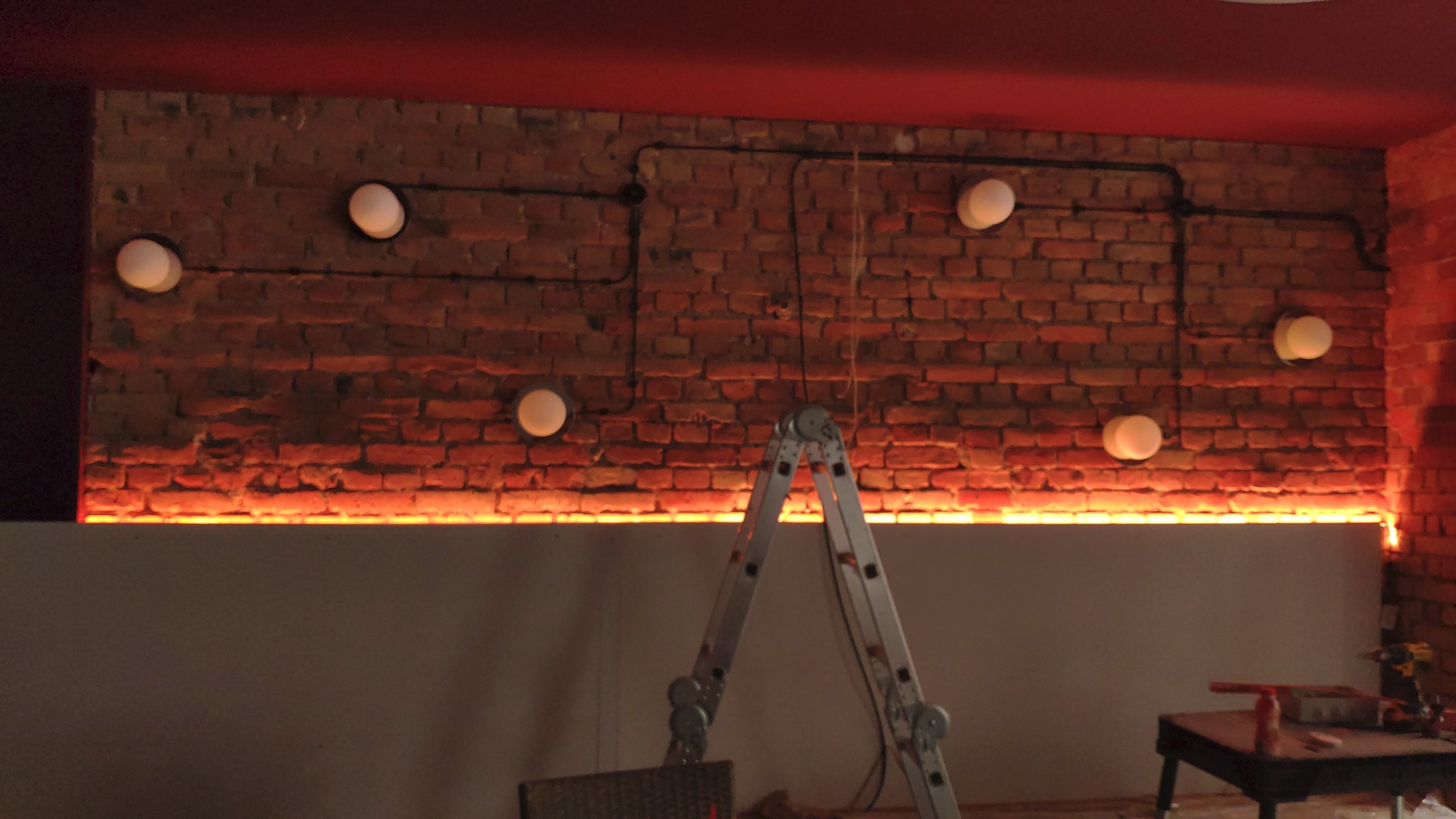 Ceglana ściana z widocznymi przewodami elektrycznymi w peszlach, okrągłe lampy na ścianie, podświetlenie LED wzdłuż krawędzi płyty gipsowej, drabina aluminiowa, czerwona poświata na suficie.