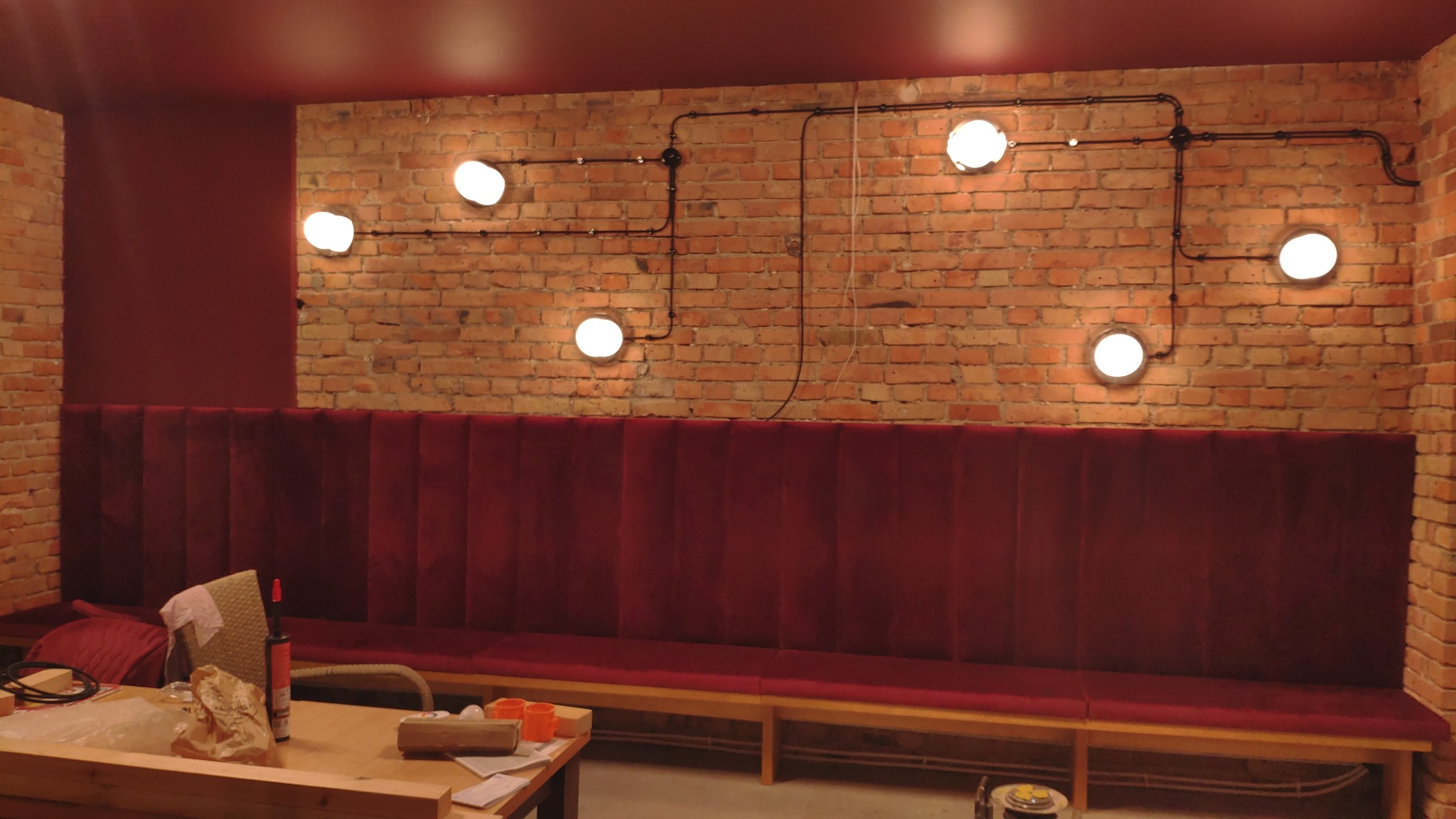 Ściana z czerwonej cegły z zamontowanymi lampami w stylu industrialnym, czarne rurki instalacyjne prowadzone po cegle, poniżej welurowa sofa w odcieniach czerwieni, na pierwszym planie drewniany...