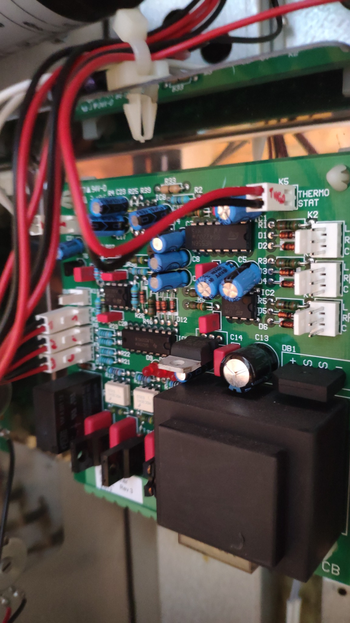 Zbliżenie na zieloną płytkę drukowaną z licznymi komponentami elektronicznymi, w tym kondensatorami, rezystorami i układami scalonymi, z widocznymi przewodami w kolorach czerwonym i czarnym.