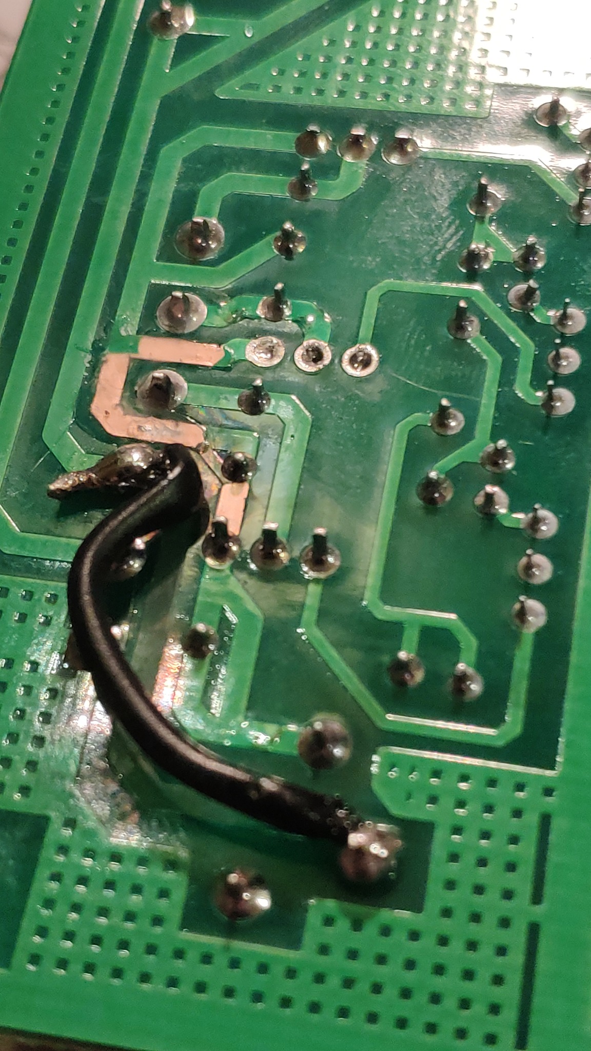 Zbliżenie na zieloną płytkę drukowaną z przylutowanym czarnym przewodem, widoczne punkty lutownicze i ścieżki przewodzące.