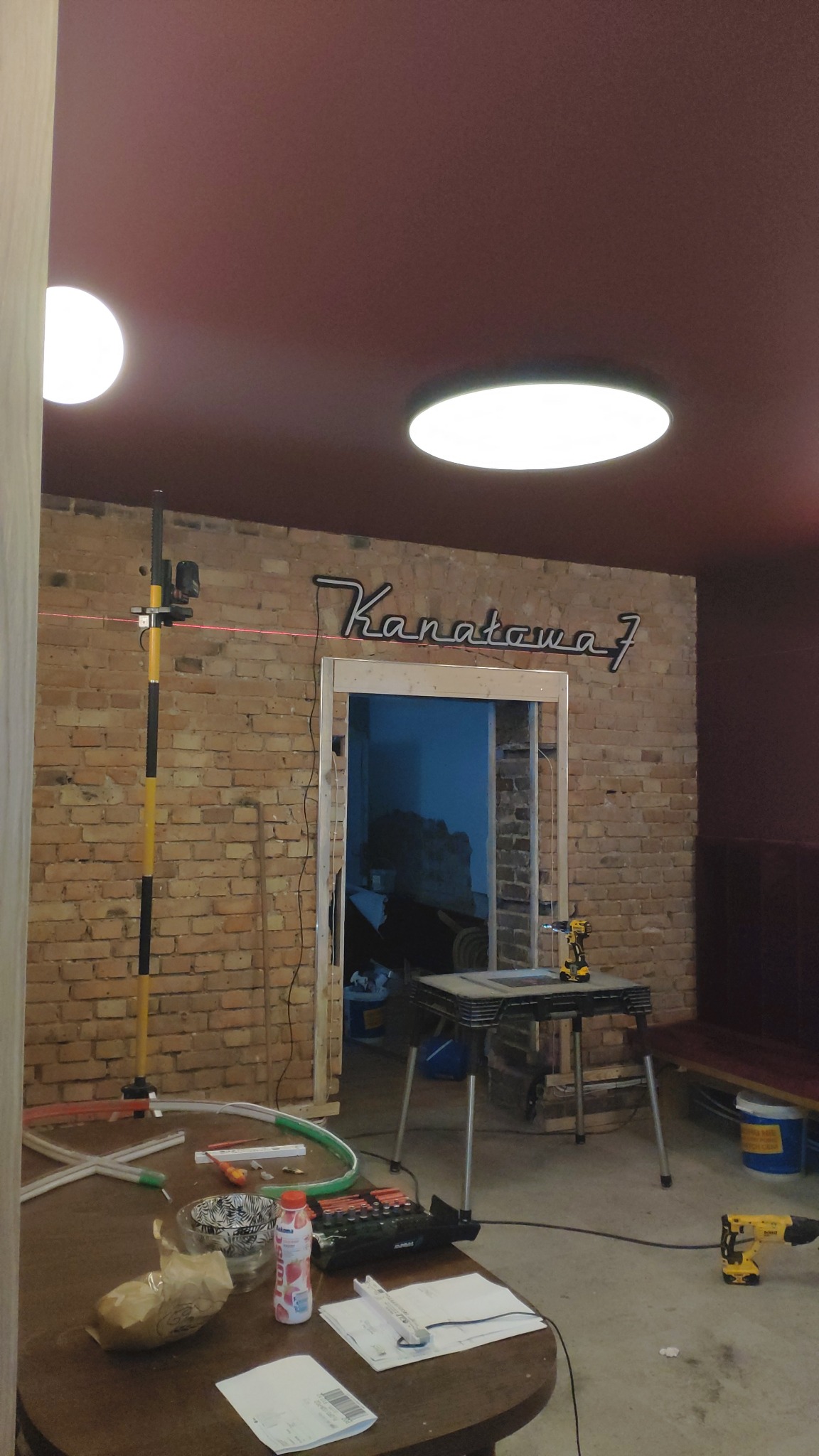 Wnętrze w trakcie remontu: ceglana ściana z wyciętym otworem, nad którym wisi podświetlany napis 'Kanatawa 7', laser krzyżowy na statywie, narzędzia i materiały budowlane na stole, sufit z dwoma...