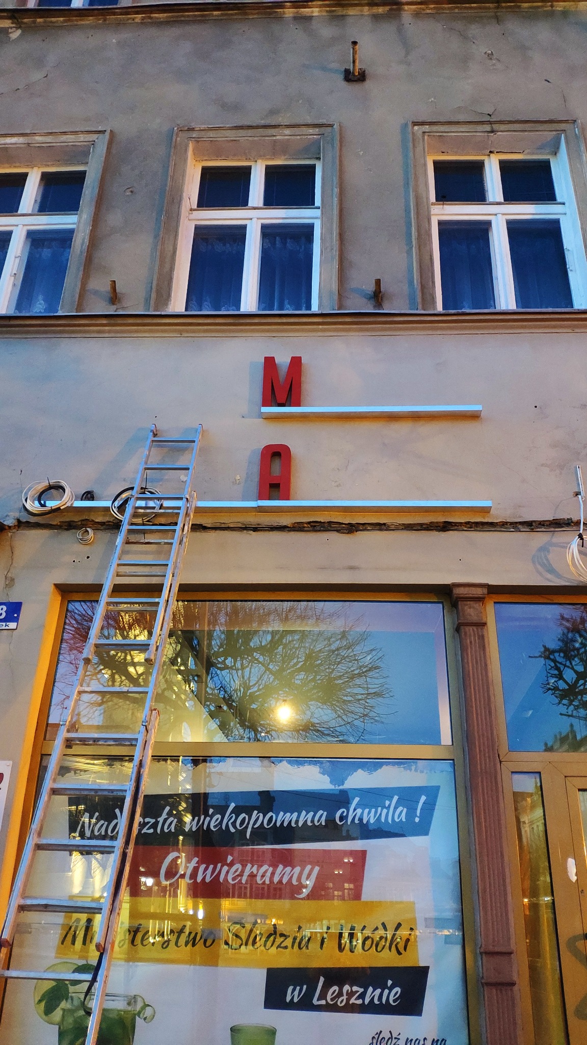 Metalowa drabina oparta o fasadę budynku z czerwonymi literami 'M' i 'A' oraz oświetleniem, widoczne okna i plakat informujący o otwarciu lokalu.