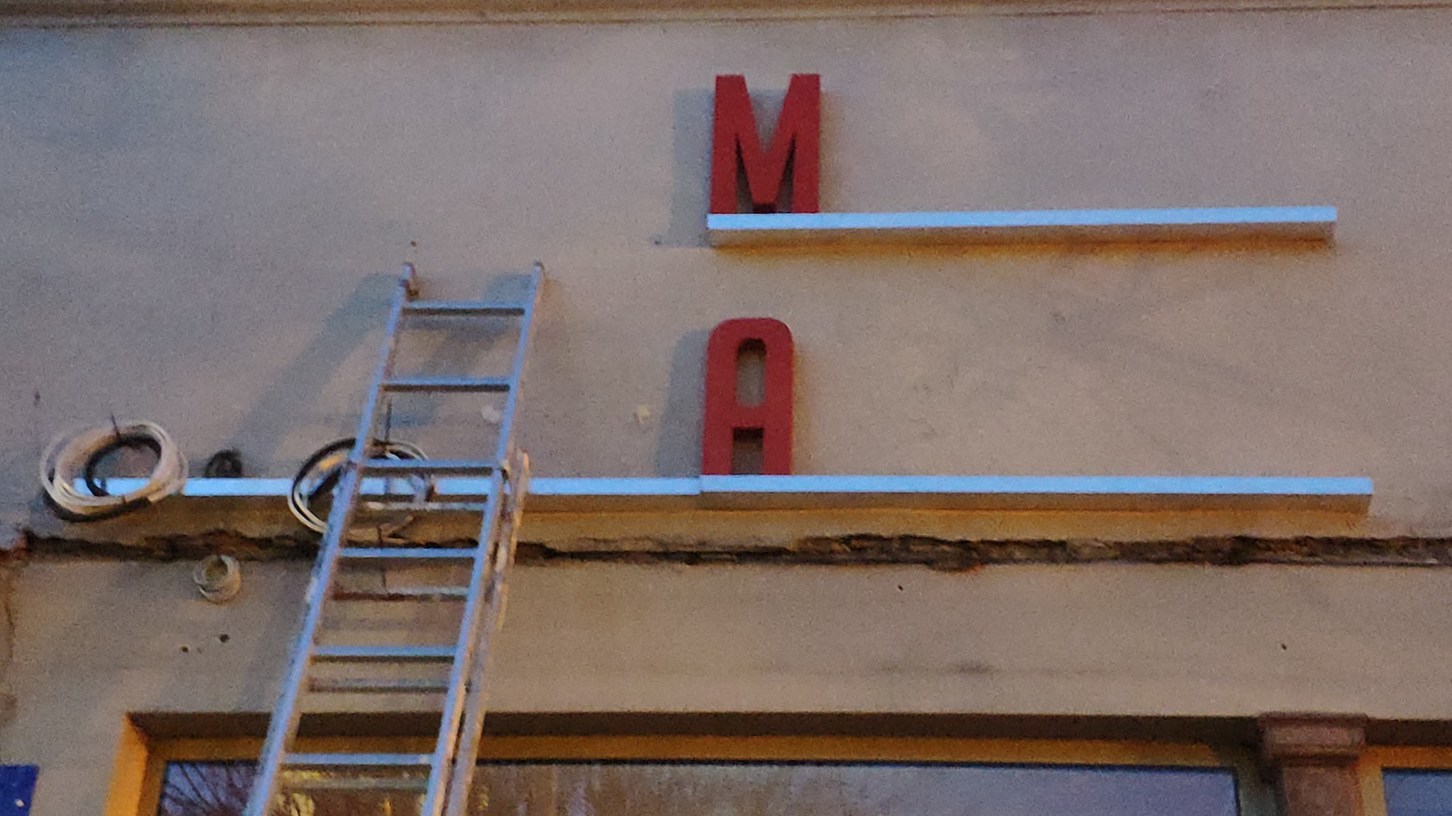 Montaż czerwonych liter 'M' i 'A' na elewacji budynku, widoczna drabina i zwój kabli elektrycznych.