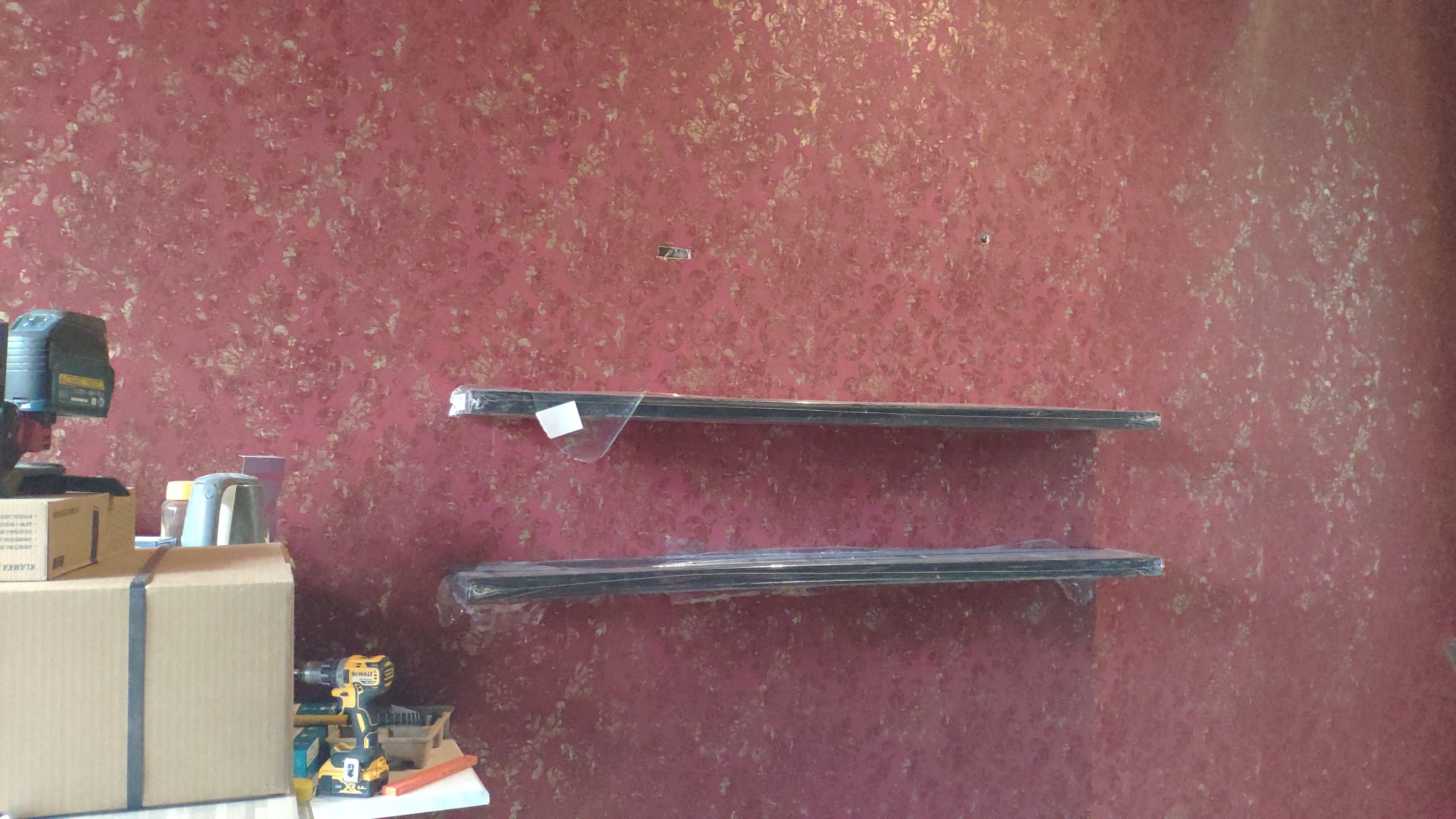 Dwie czarne półki, jeszcze w folii, przygotowane do montażu na czerwonej ścianie o kwiecistym wzorze. Na pierwszym planie narzędzia: wiertarka akumulatorowa i skrzynka z akcesoriami, sugerujące...