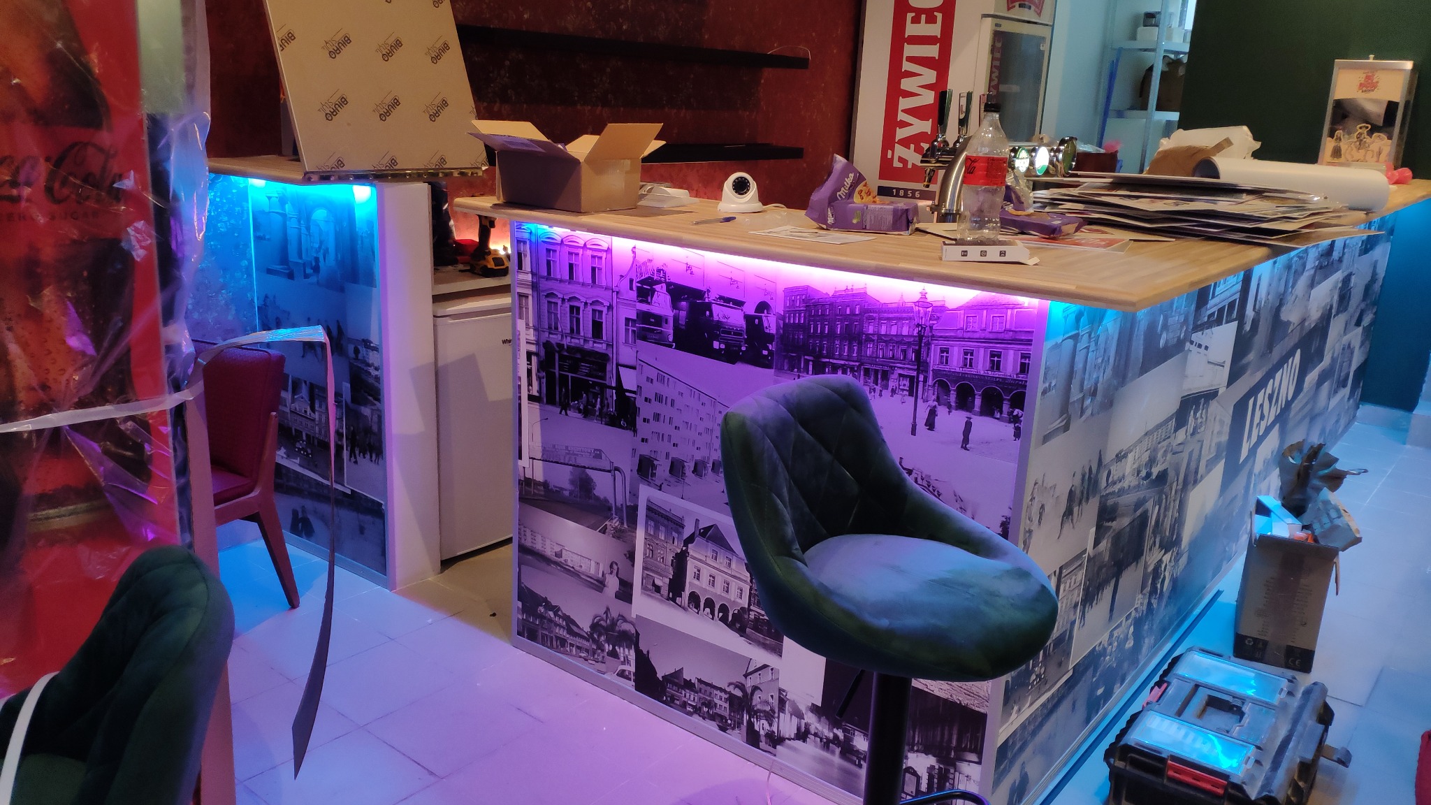 Bar z podświetleniem LED, oklejony fototapetą z motywem starego miasta, krzesło barowe, narzędzia i materiały budowlane w trakcie prac wykończeniowych.
