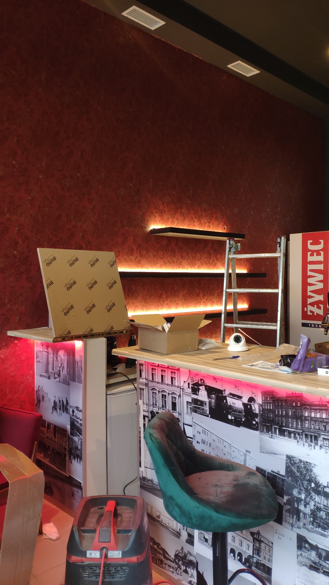 Montaż oświetlenia LED na ścianie w pomieszczeniu z elementami dekoracyjnymi, widoczna drabina, narzędzia i materiały montażowe.