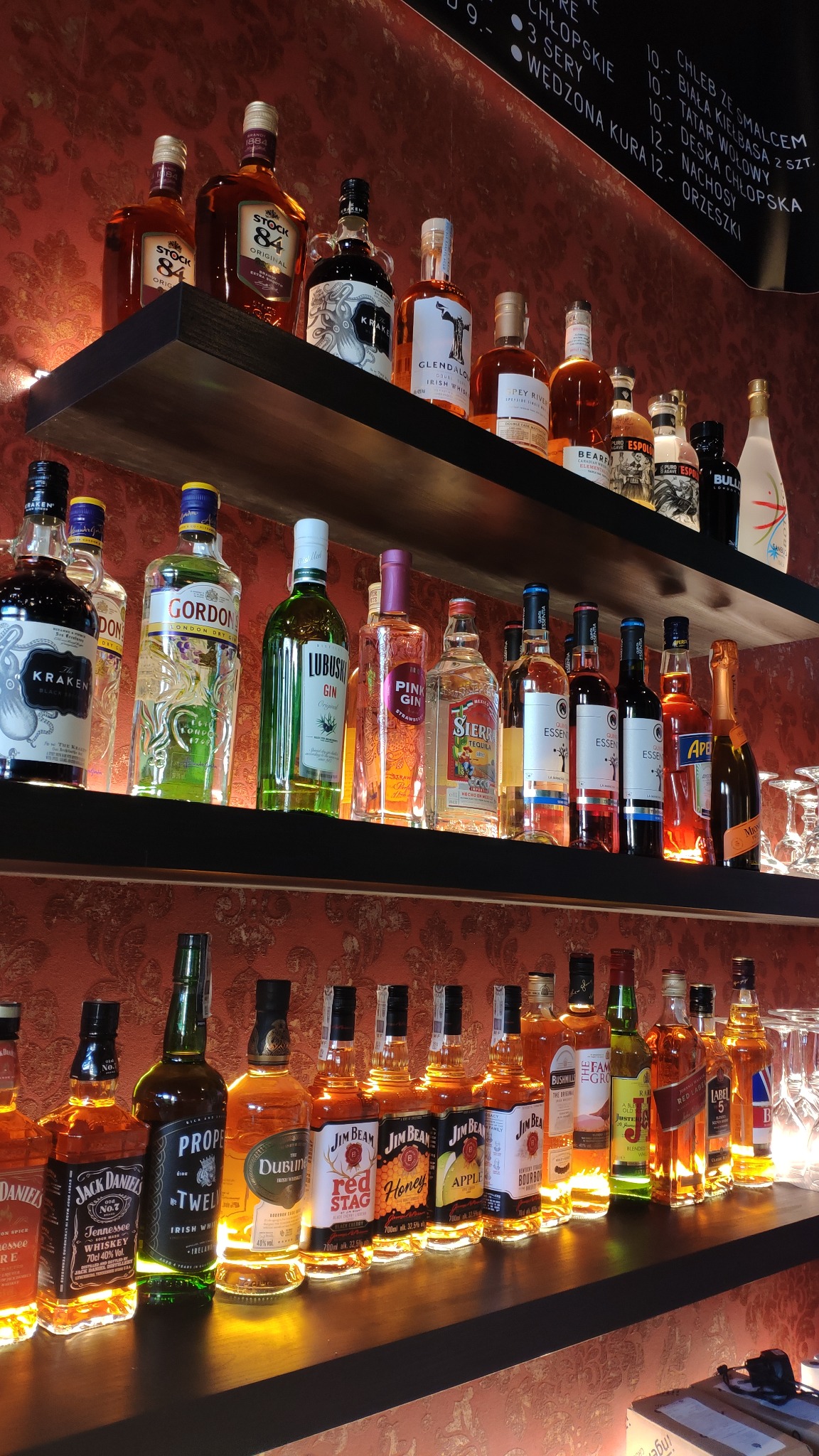 Półki barowe z podświetleniem, pełne różnorodnych butelek alkoholi, w tym whisky Jack Daniel's i Jim Beam, gin Gordon's i Lubuski, tequila Sierra, z dekoracyjną tapetą w tle i menu lokalu widocznym...