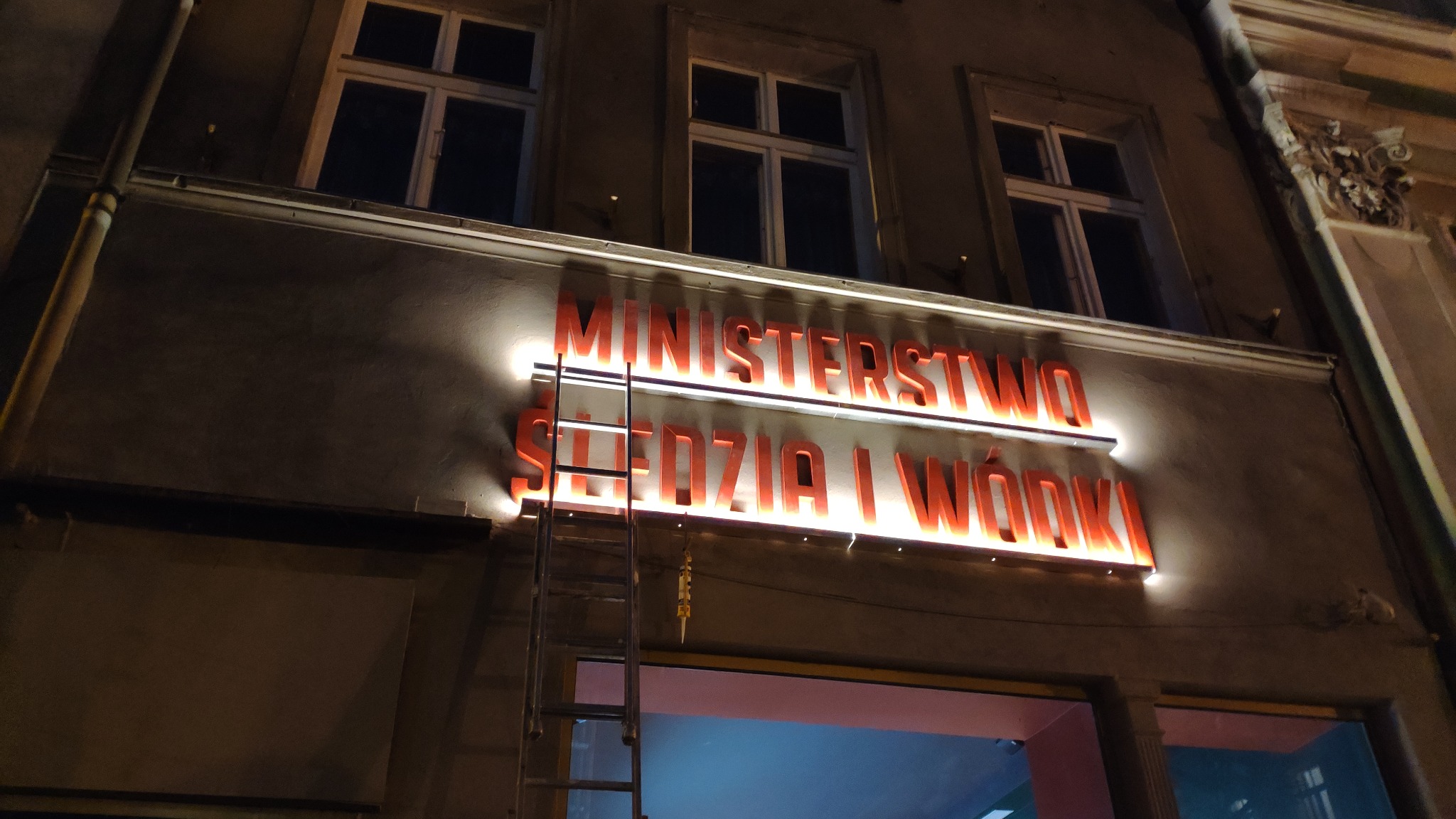 Montaż podświetlanego szyldu 'Ministerstwo Śledzia i Wódki' na fasadzie budynku w Poznaniu, widoczna drabina oparta o budynek.