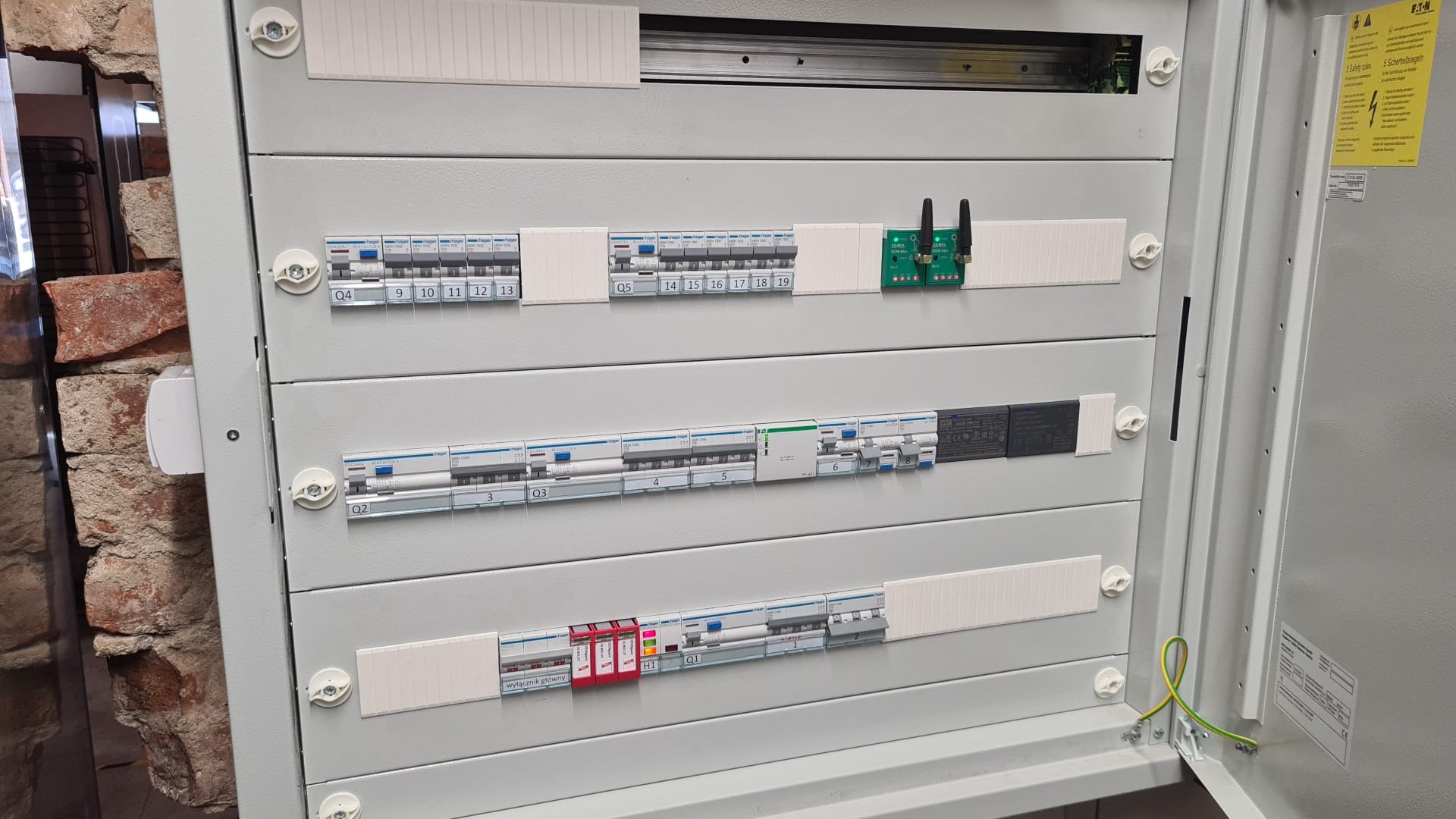 Otwarta skrzynka rozdzielcza z bezpiecznikami, wyłącznikami i modułem sterującym z antenami, zamontowana przy ceglanej ścianie.