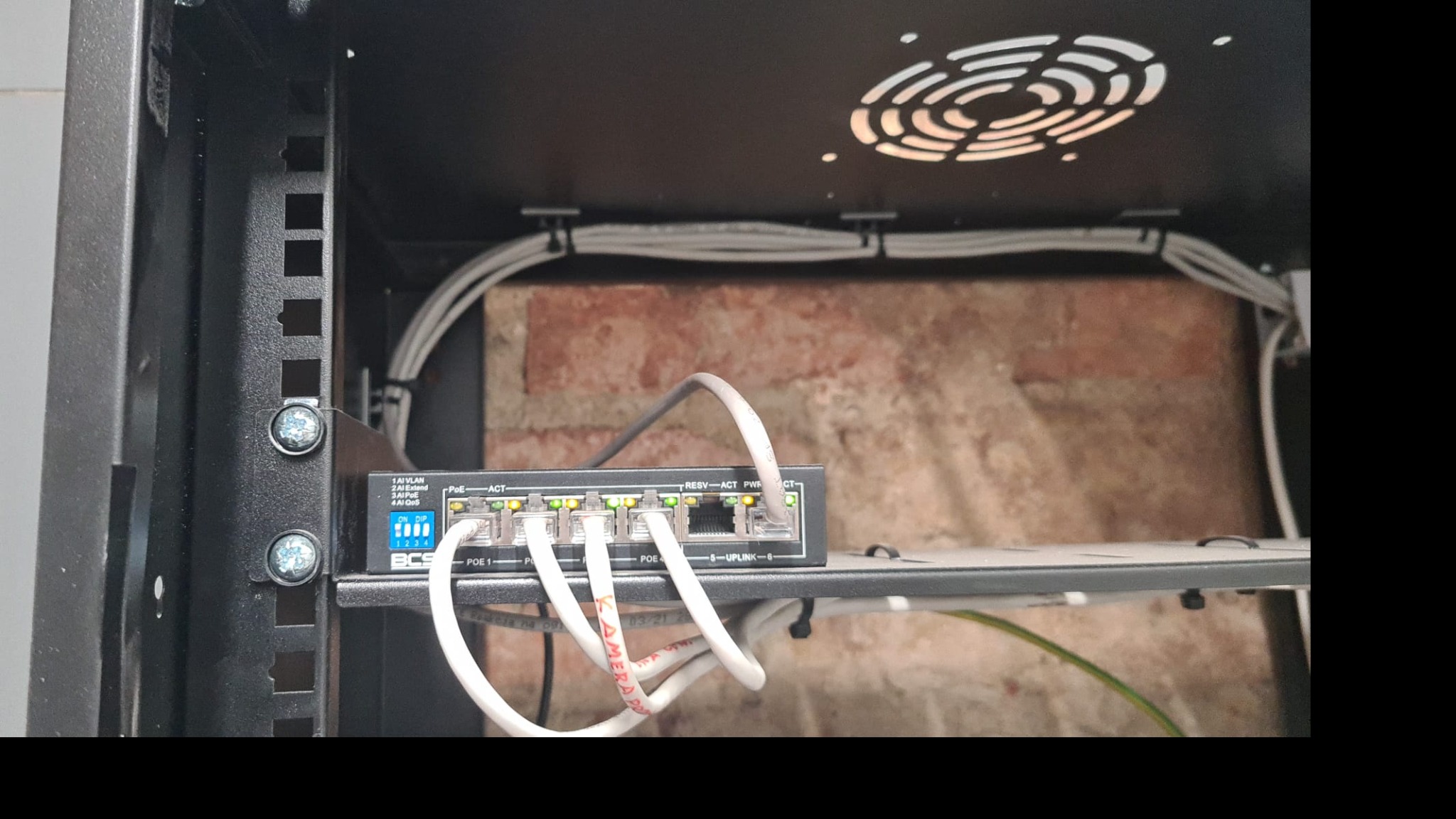 Switch sieciowy BCS zamontowany w szafie rack, z podłączonymi kablami Ethernet, widoczne diody sygnalizujące pracę portów PoE, kable ułożone w korytkach, ściana z cegły w tle.