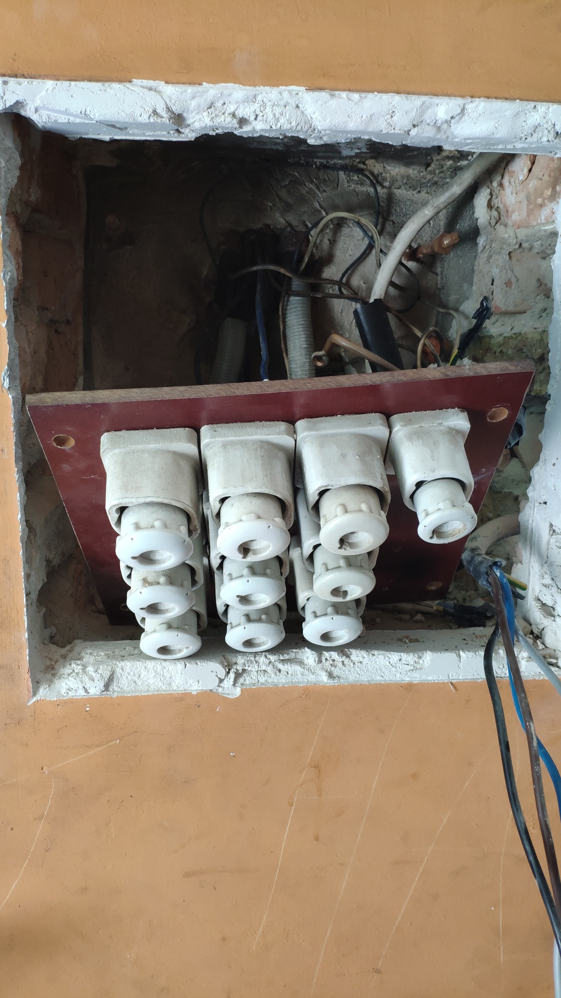 Otwór w ścianie z widoczną tablicą z ceramicznymi bezpiecznikami topikowymi, okablowaniem i fragmentami muru.