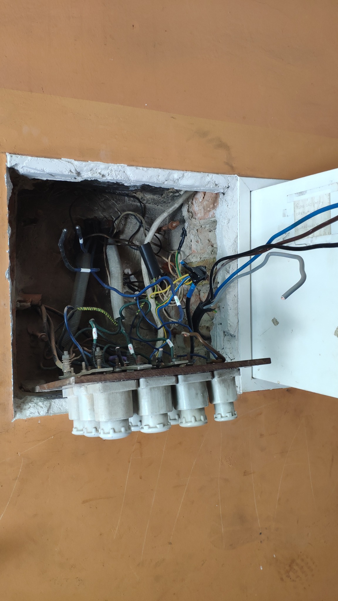 Otwarta skrzynka z bezpiecznikami topikowymi w ścianie, widoczne okablowanie i instalacja elektryczna w starej kamienicy podczas wymiany zabezpieczeń.