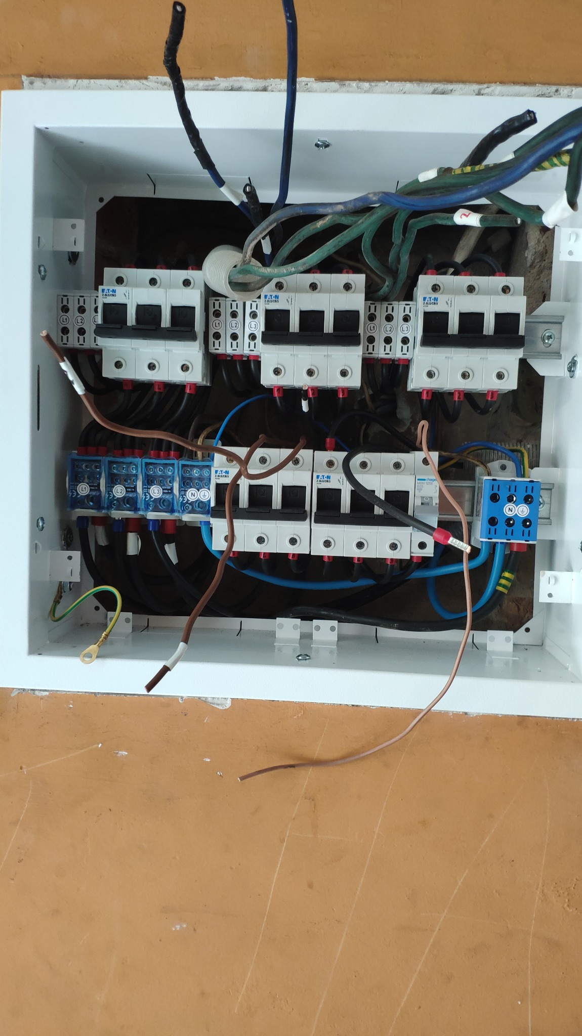 Otwarta skrzynka elektryczna z zamontowanymi bezpiecznikami Eaton i okablowaniem o różnych kolorach, widoczne przewody miedziane z końcówkami oczkowymi i ślady po starych instalacjach na ścianie.