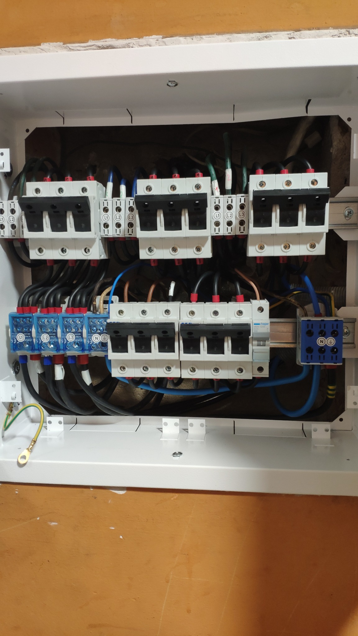 Wnętrze białej skrzynki rozdzielczej z zamontowanymi bezpiecznikami i przewodami, widoczne niebieskie i czarne kable, złącze uziemiające z żółto-zielonym przewodem, oraz oznaczenia L1, L2, L3...