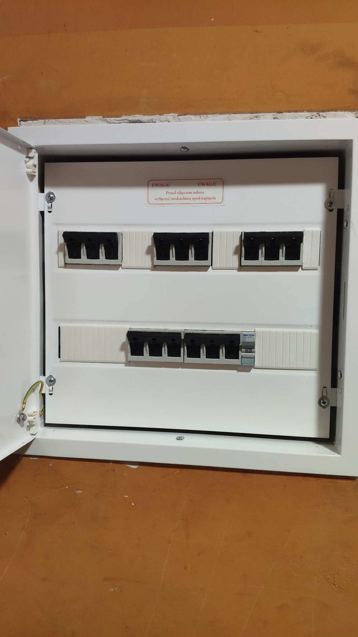 Otwarta skrzynka bezpiecznikowa z widocznymi bezpiecznikami i naklejką ostrzegawczą 'Przed zdjęciem osłony wyłączyć rozdzielnicę spod napięcia'.