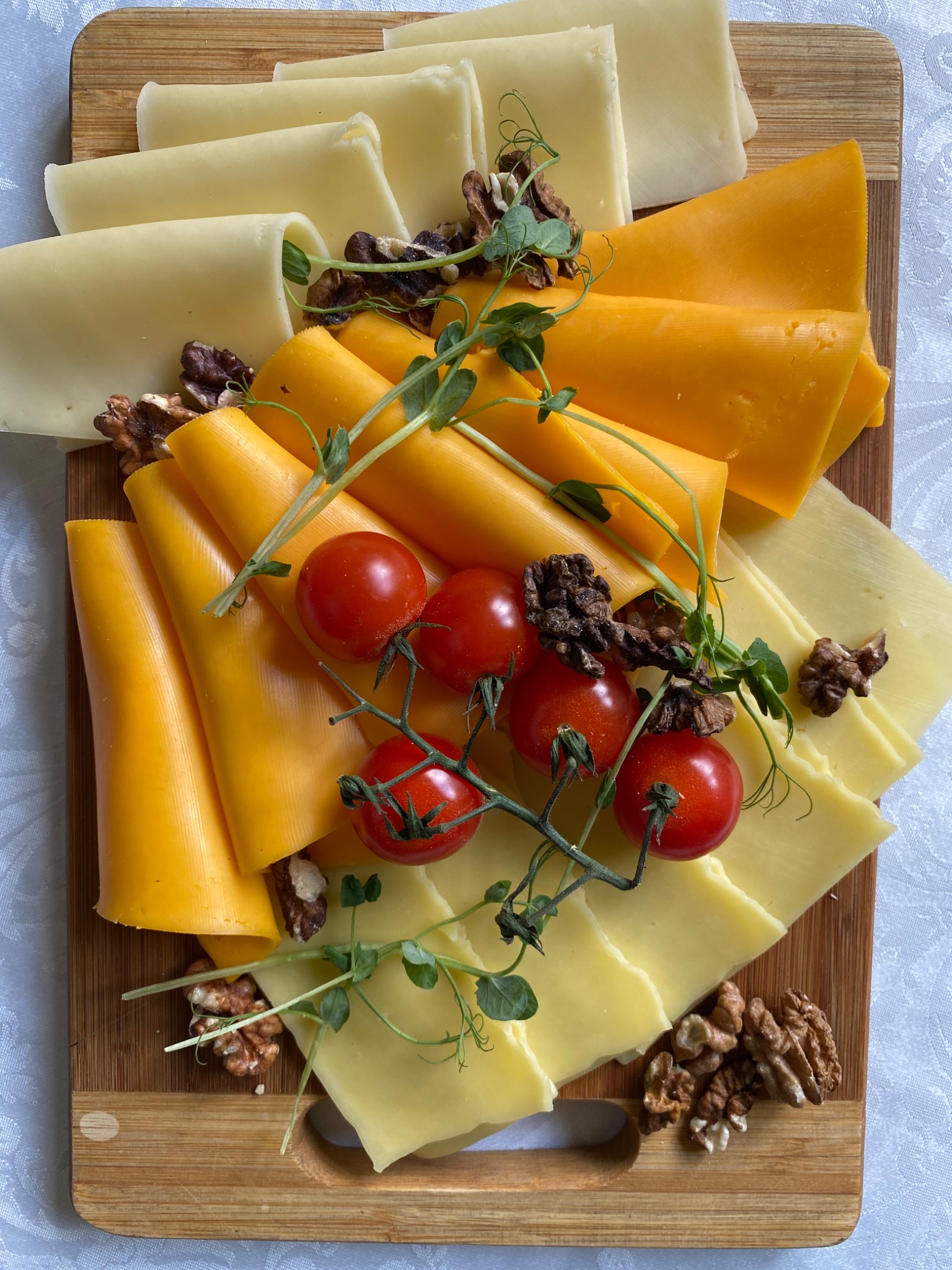 Deska serów z dwoma rodzajami sera, udekorowana gałązką pomidorków koktajlowych, orzechami włoskimi i mikro-ziołami na białym obrusie.