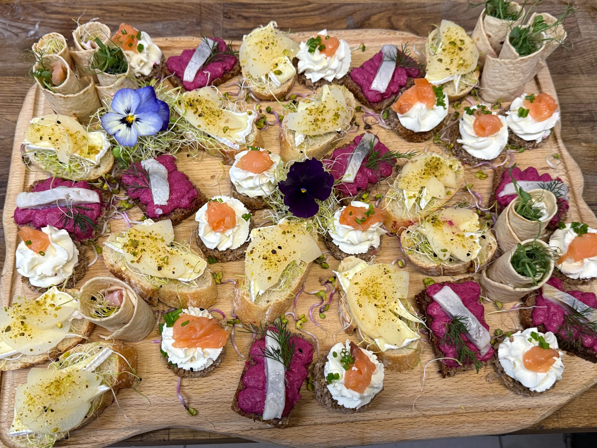 Drewniana deska z różnorodnymi, artystycznie ułożonymi kanapeczkami, dekorowanymi kwiatami i świeżymi ziołami. Widoczne kompozycje z łososiem, śledziem i kremowym serem.