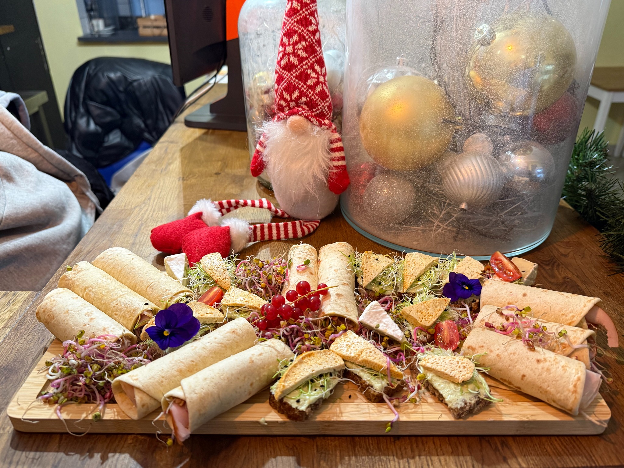 Świąteczna deska przekąsek: tortille, kanapki, ser, dekoracje. Udekorowana kwiatami i kiełkami, obok skrzat i bombki w szklanym naczyniu. Drewniany stół.