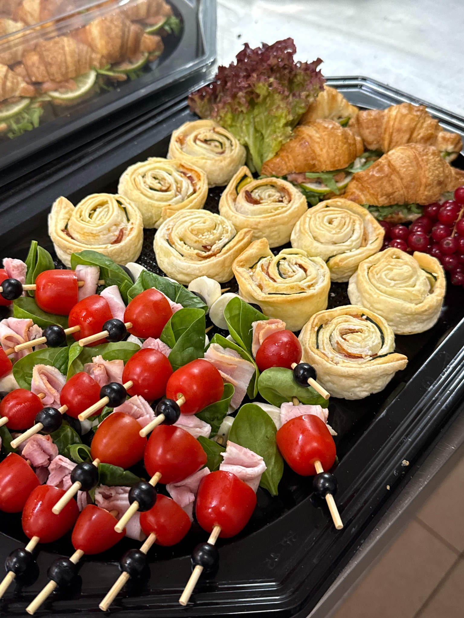 Elegancka taca cateringowa z roladkami z ciasta francuskiego, mini-szaszłykami z pomidorków i szynki, oraz mini-kanapeczkami z croissantów, dekorowana sałatą i czerwoną porzeczką.