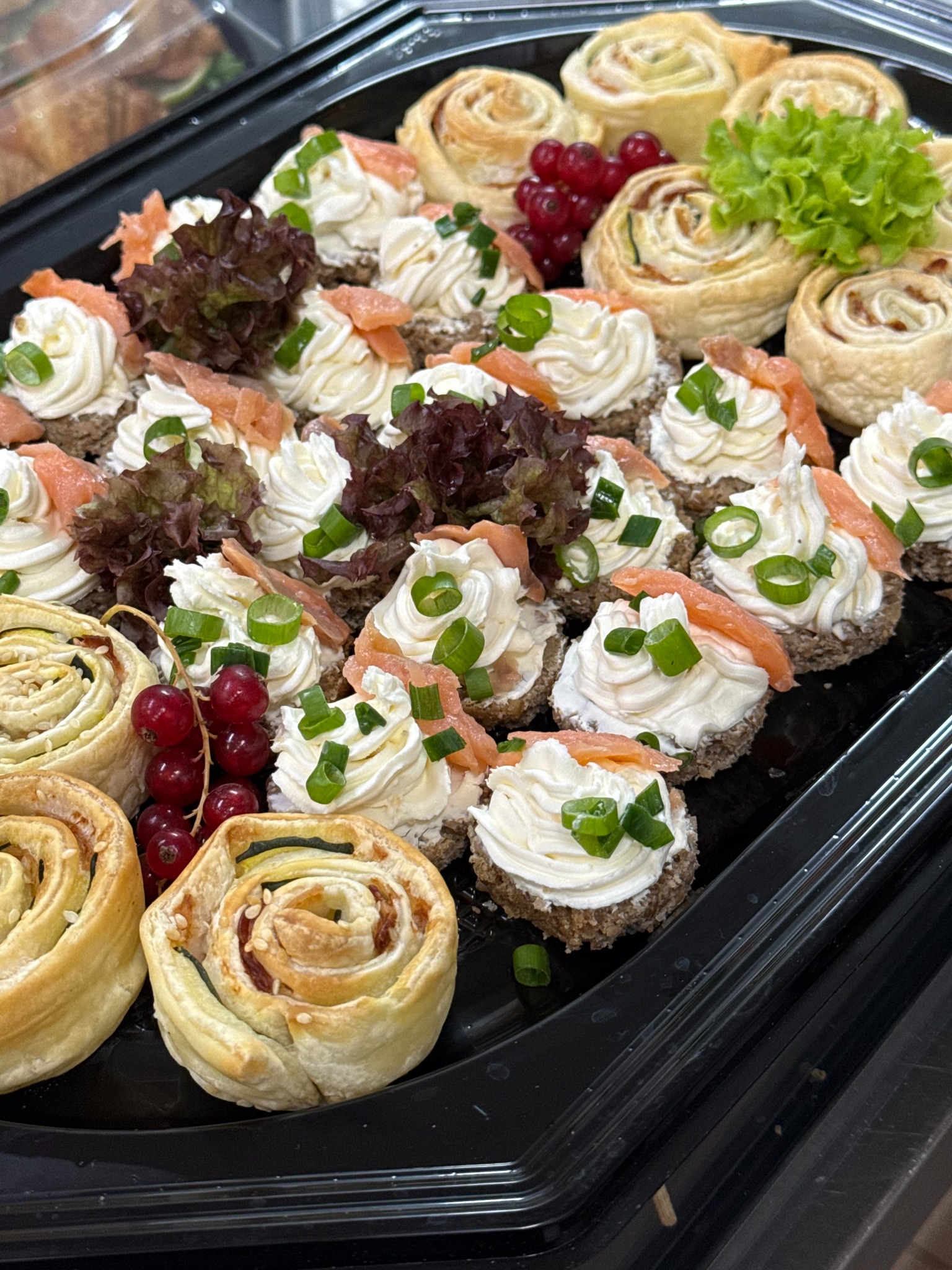 Elegancka taca z wykwintnymi przekąskami: roladki z ciasta francuskiego i kanapeczki z kremowym serkiem, łososiem i szczypiorkiem, udekorowane czerwoną porzeczką i sałatą.