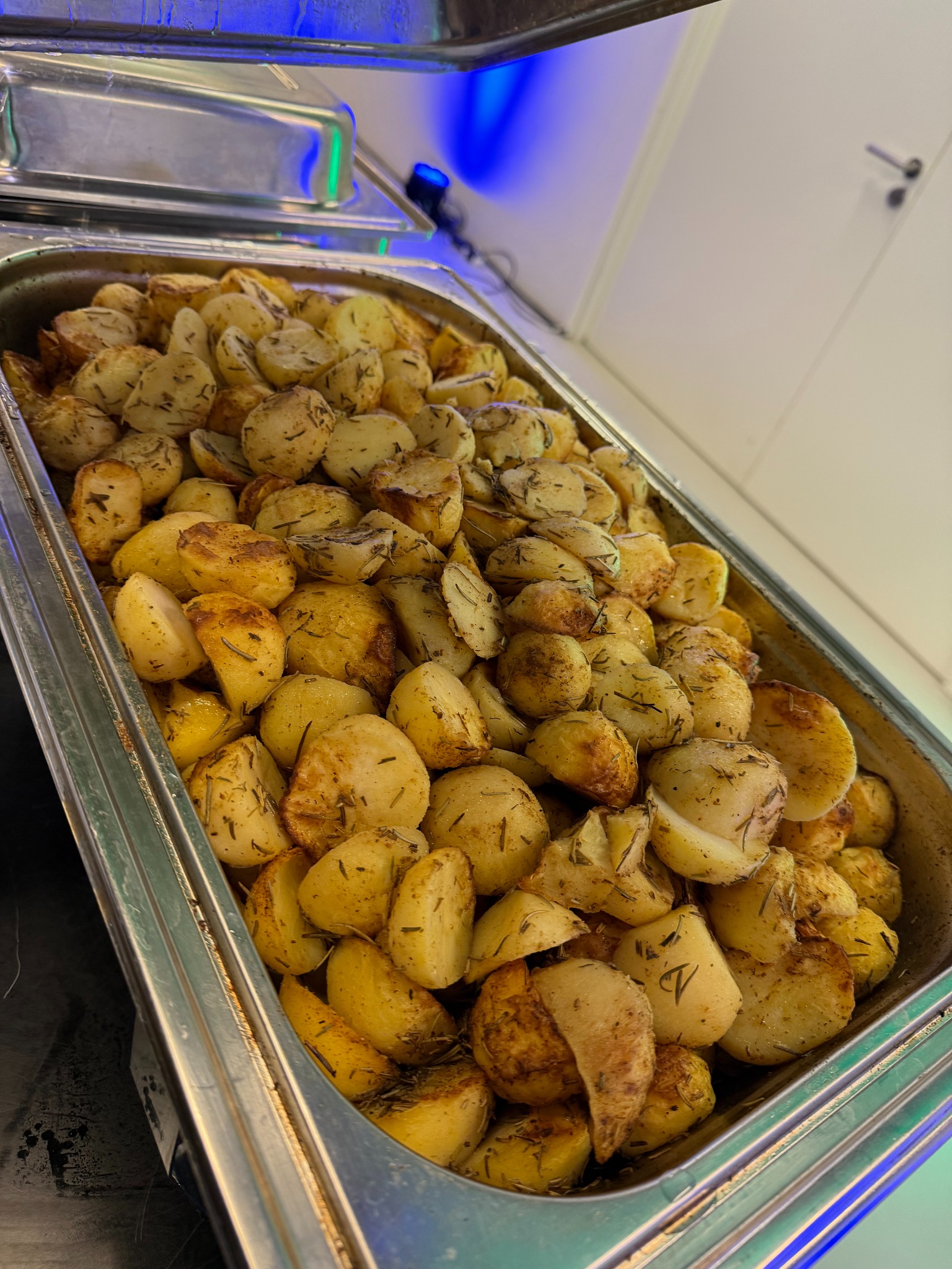 Metalowa patera wypełniona pieczonymi ziemniakami z ziołami, oświetlona niebieskim światłem, gotowa do podania na przyjęciu cateringowym.