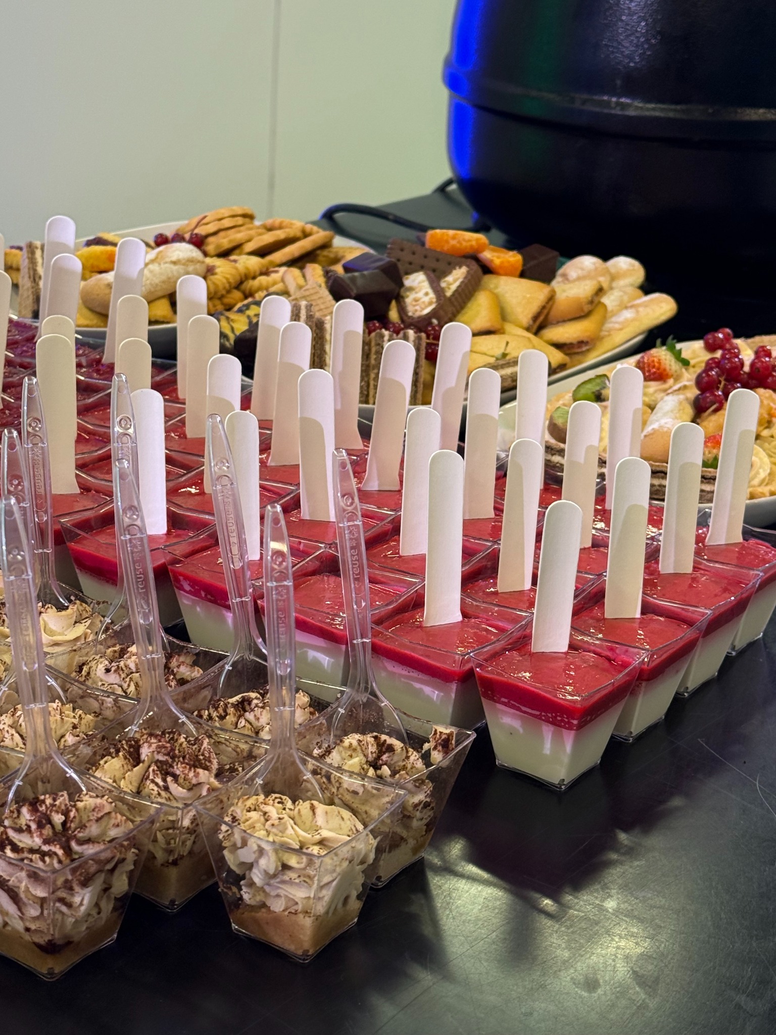 Desery w plastikowych pojemnikach: tiramisu z posypką kakaową, panna cotta z musem malinowym i drewnianymi patyczkami, ciastka na paterach w tle.