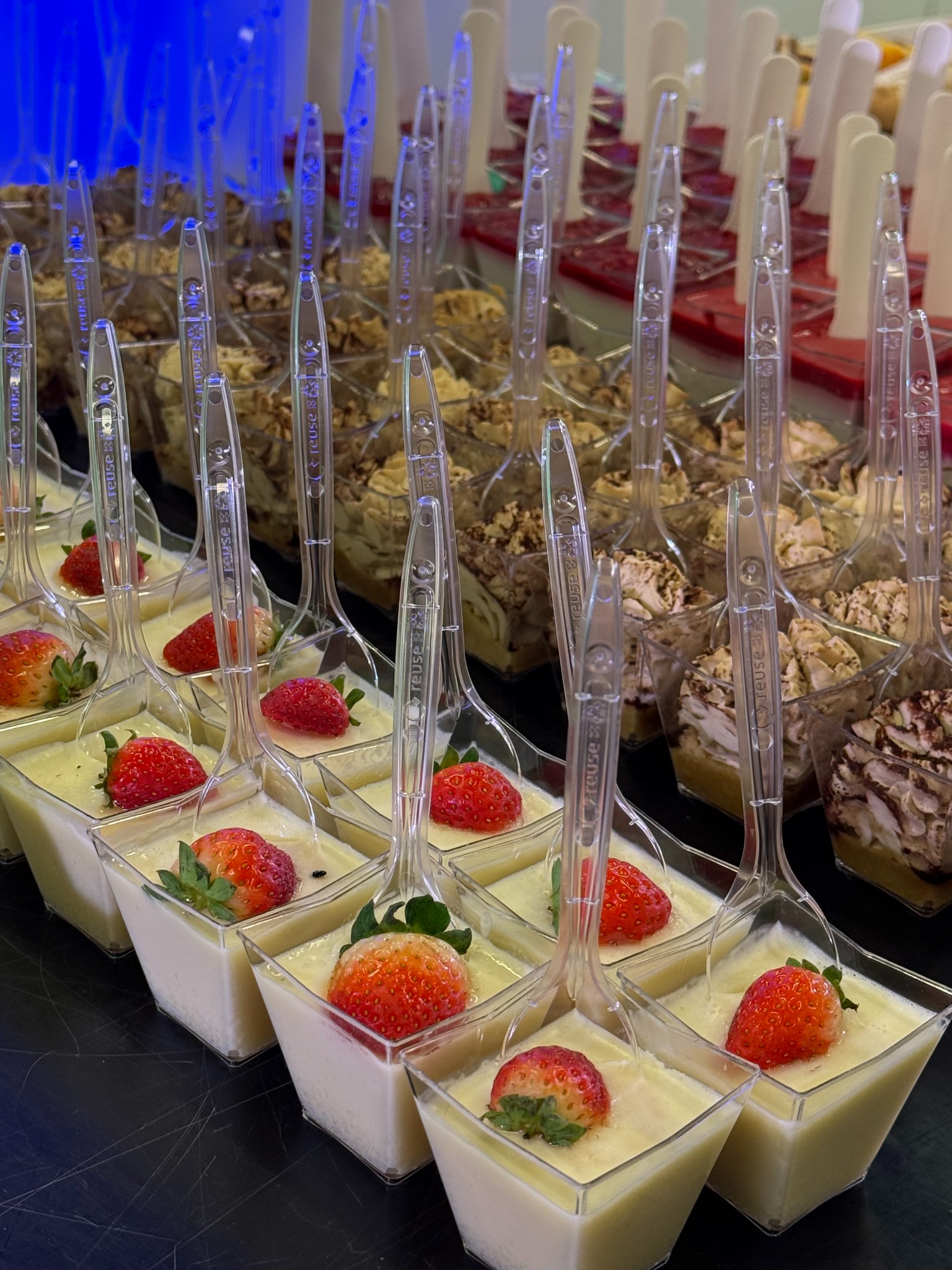 Rzędy deserów w kwadratowych naczyniach z łyżeczkami, część z kremem i truskawkami, część z posypką, ustawione na ciemnym blacie pod niebieskim światłem.