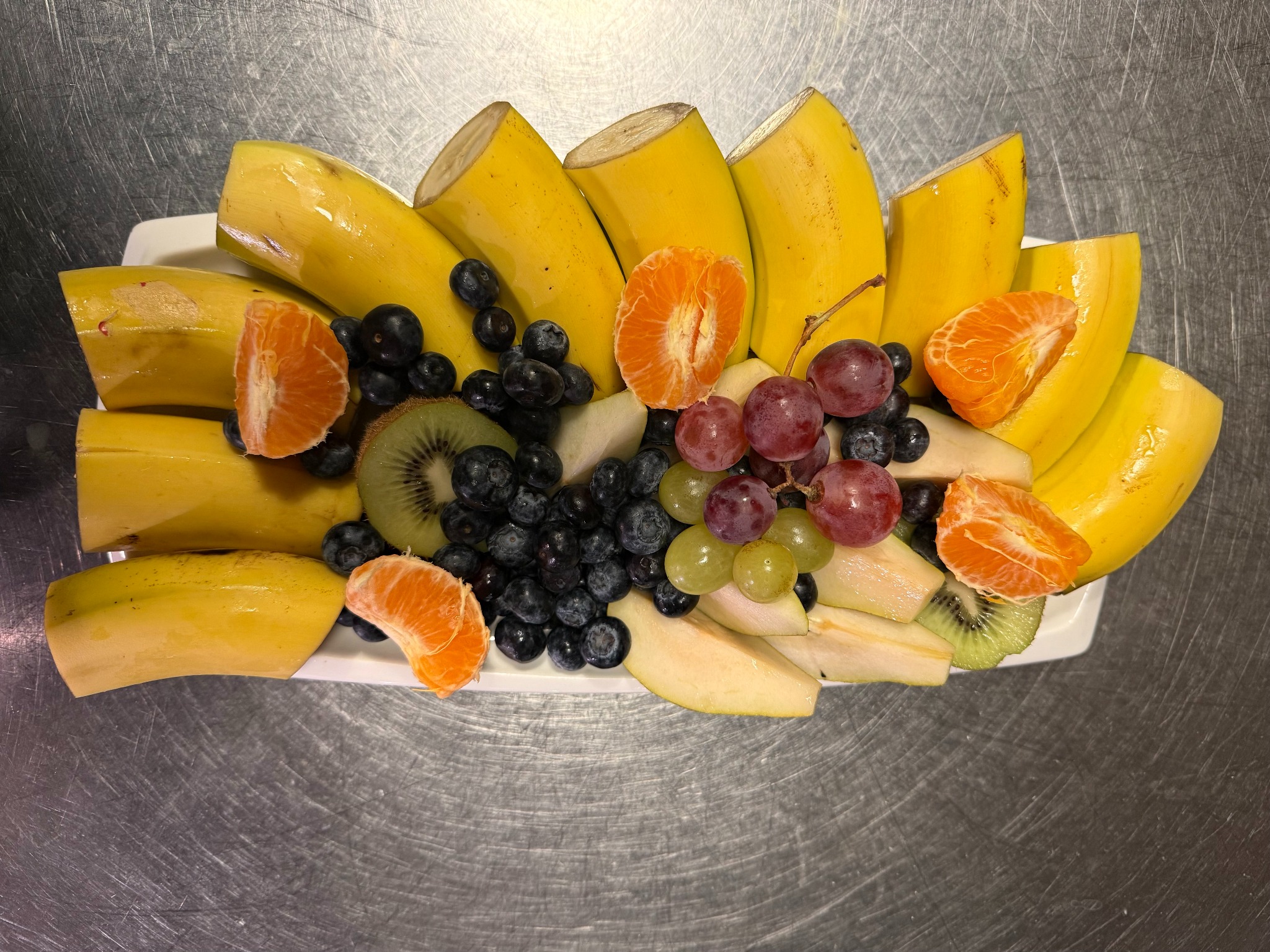 Dekoracyjna taca z pokrojonymi bananami, mandarynkami, kiwi, winogronami i borówkami na metalowym blacie. Owocowa kompozycja cateringowa.