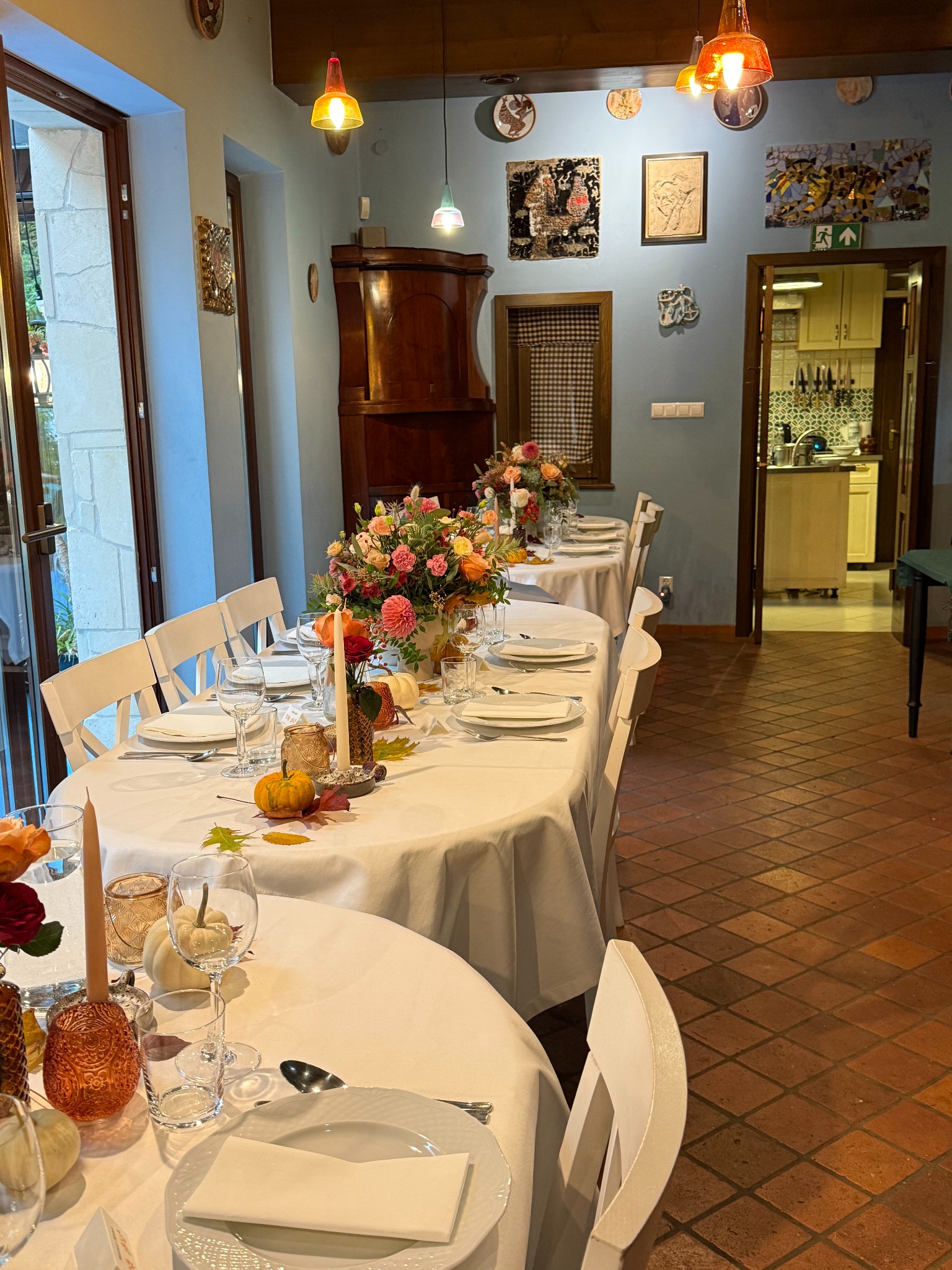 Uroczysty stół z białym obrusem, kwiatami, dyniami i świecami. Dekoracja jesienna, zastawa stołowa. Widok na kuchnię w tle. Przytulne wnętrze restauracji.