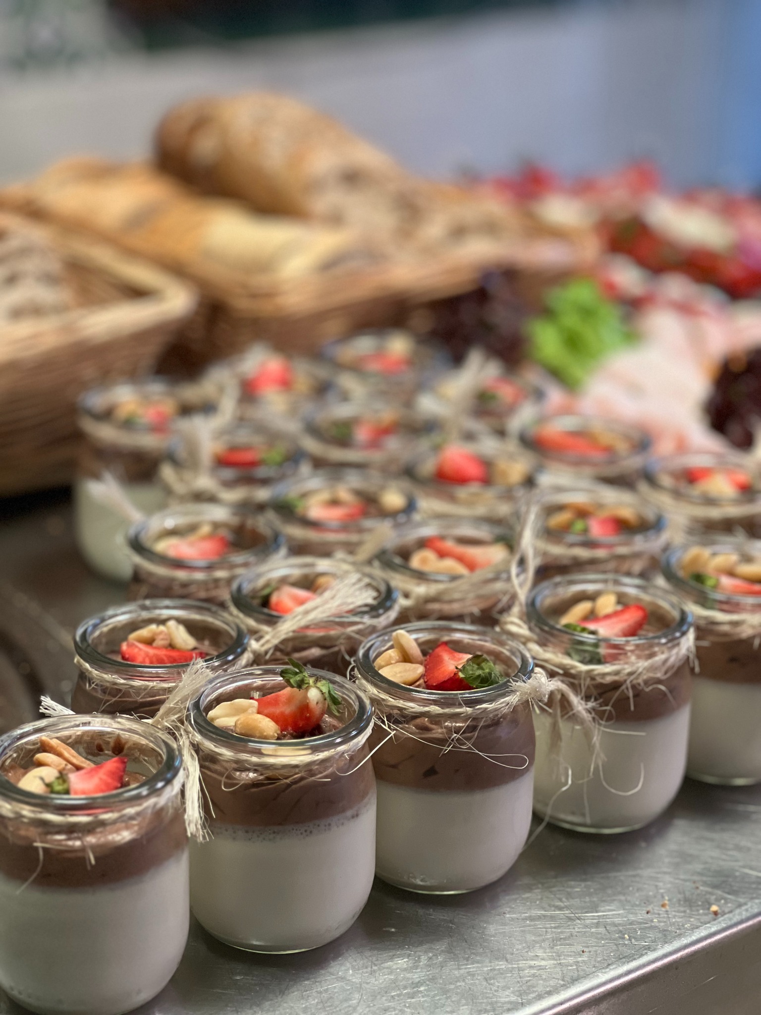 Deser w słoiczkach z truskawkami i orzechami, ustawiony na metalowej powierzchni. W tle pieczywo i wędliny. Elegancki catering na przyjęcie.