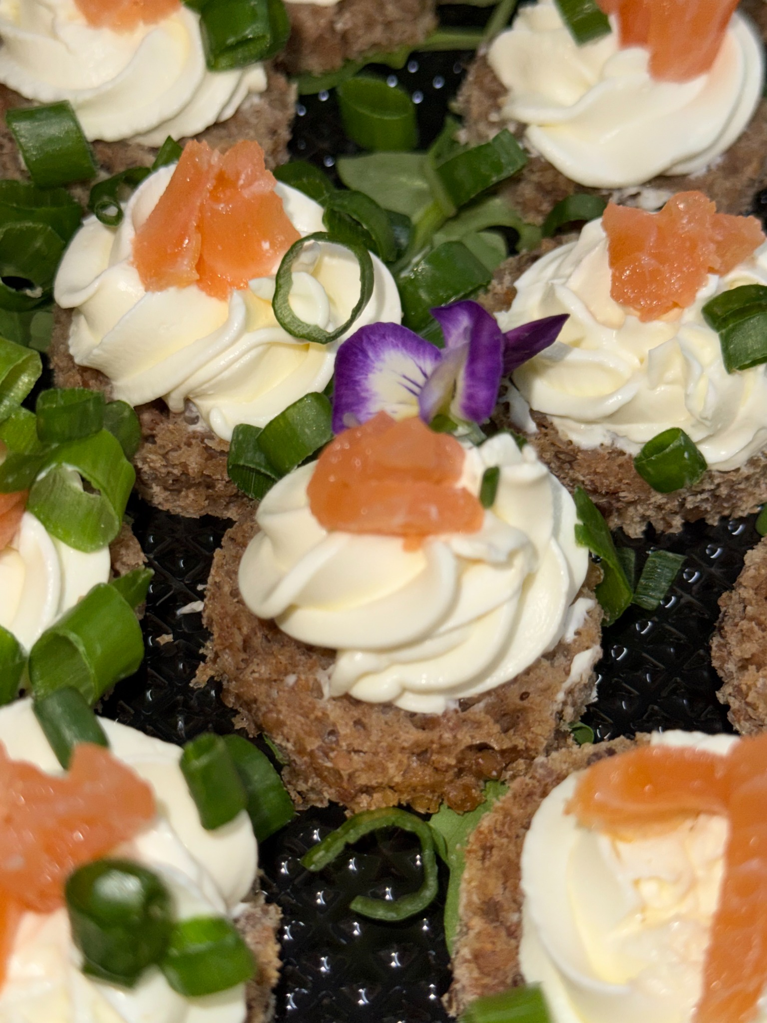 Apetyczne kanapeczki na ciemnym pieczywie, udekorowane kremowym serkiem, łososiem, szczypiorkiem i fioletowym kwiatkiem, prezentowane z bliska na czarnym talerzu.