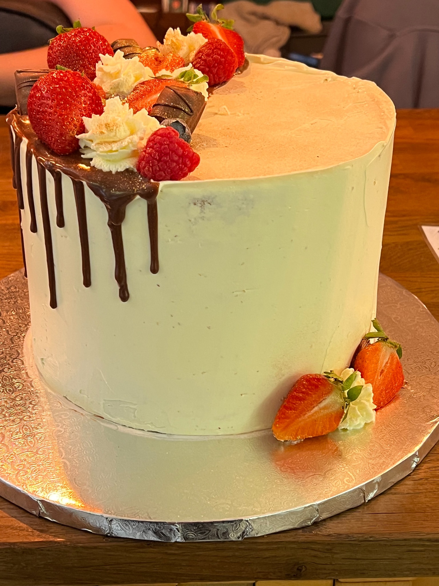 Tort z kremem, polany czekoladą i udekorowany świeżymi truskawkami, malinami i kawałkami czekolady, na srebrnej paterze. Widoczne detale dekoracji i tekstura kremu.