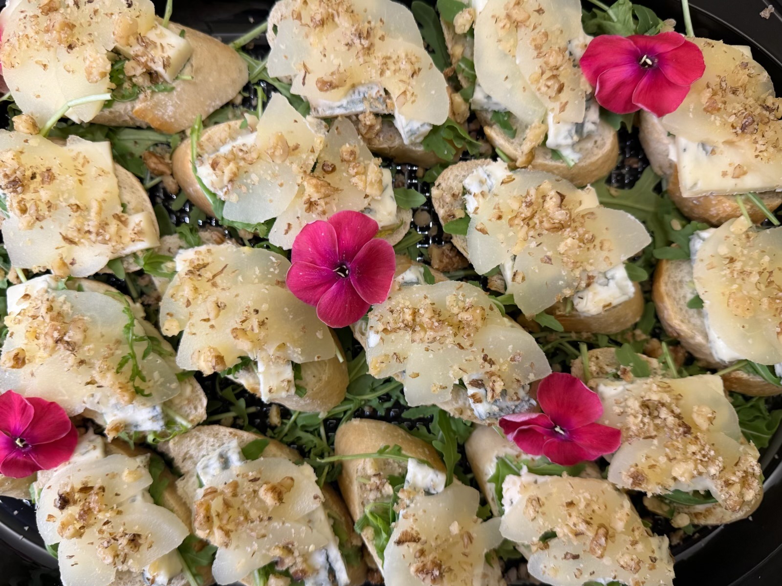 Dekoracyjne kanapeczki z serem pleśniowym, gruszką, orzechami i rukolą, ozdobione jadalnymi kwiatami na czarnej tacy. Elegancki catering na imprezę.