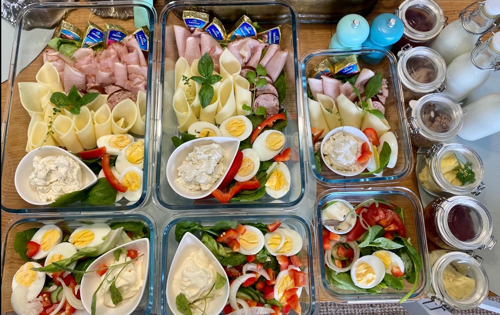Zestaw lunchboxów z różnorodnymi przekąskami: wędliny, sery, jajka, warzywa i sosy. Estetyczne i zdrowe posiłki w szklanych pojemnikach, idealne na catering.