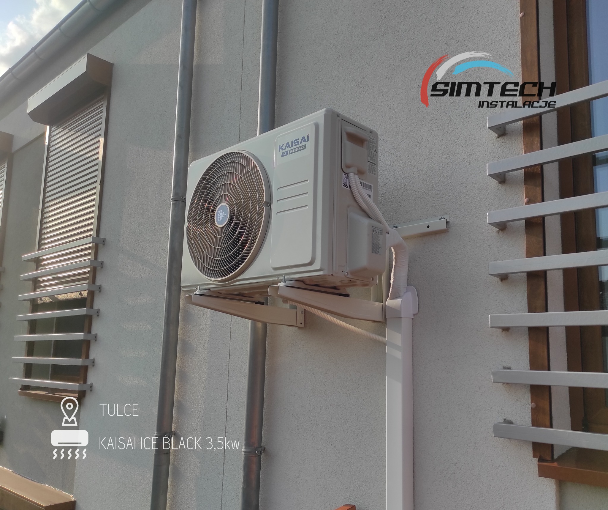 Zewnętrzna jednostka klimatyzacji Kaisai Ice Black 3,5kW zamontowana na elewacji budynku, widoczna rura spustowa i fragment okna z żaluzjami zewnętrznymi, logo firmy Simtech Instalacje w prawym...