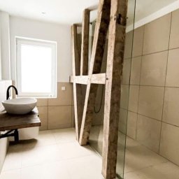 Bolo bud - Nowoczesna łazienka z kabiną prysznicową oddzieloną szklaną ścianą wspartą na drewnianych belkach, umywalka nablatowa na drewnianym blacie, jasne płytki na ścianach i podłodze, minimalistyczny...