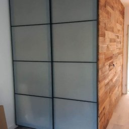 Bolo bud - Szklana szafa przesuwna z czarnymi ramami, sąsiadująca z drewnianą ścianą z desek w pomieszczeniu z szarymi panelami podłogowymi.