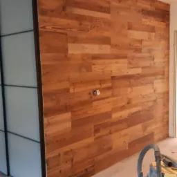 Ściana wykończona panelami z naturalnego drewna o ciepłym odcieniu, z widocznymi gniazdkami elektrycznymi i fragmentem szafy z przesuwnymi drzwiami po lewej stronie. Na podłodze widoczny sprzęt...