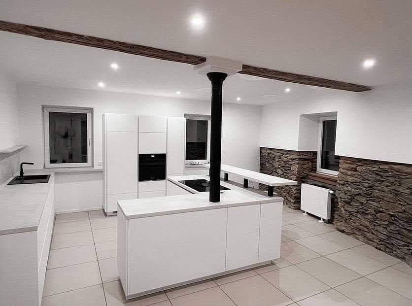 Nowoczesna, minimalistyczna kuchnia z białą zabudową i wyspą kuchenną, akcentowana drewnianymi belkami stropowymi i fragmentami ściany wykończonymi kamieniem dekoracyjnym. Czarna kolumna...