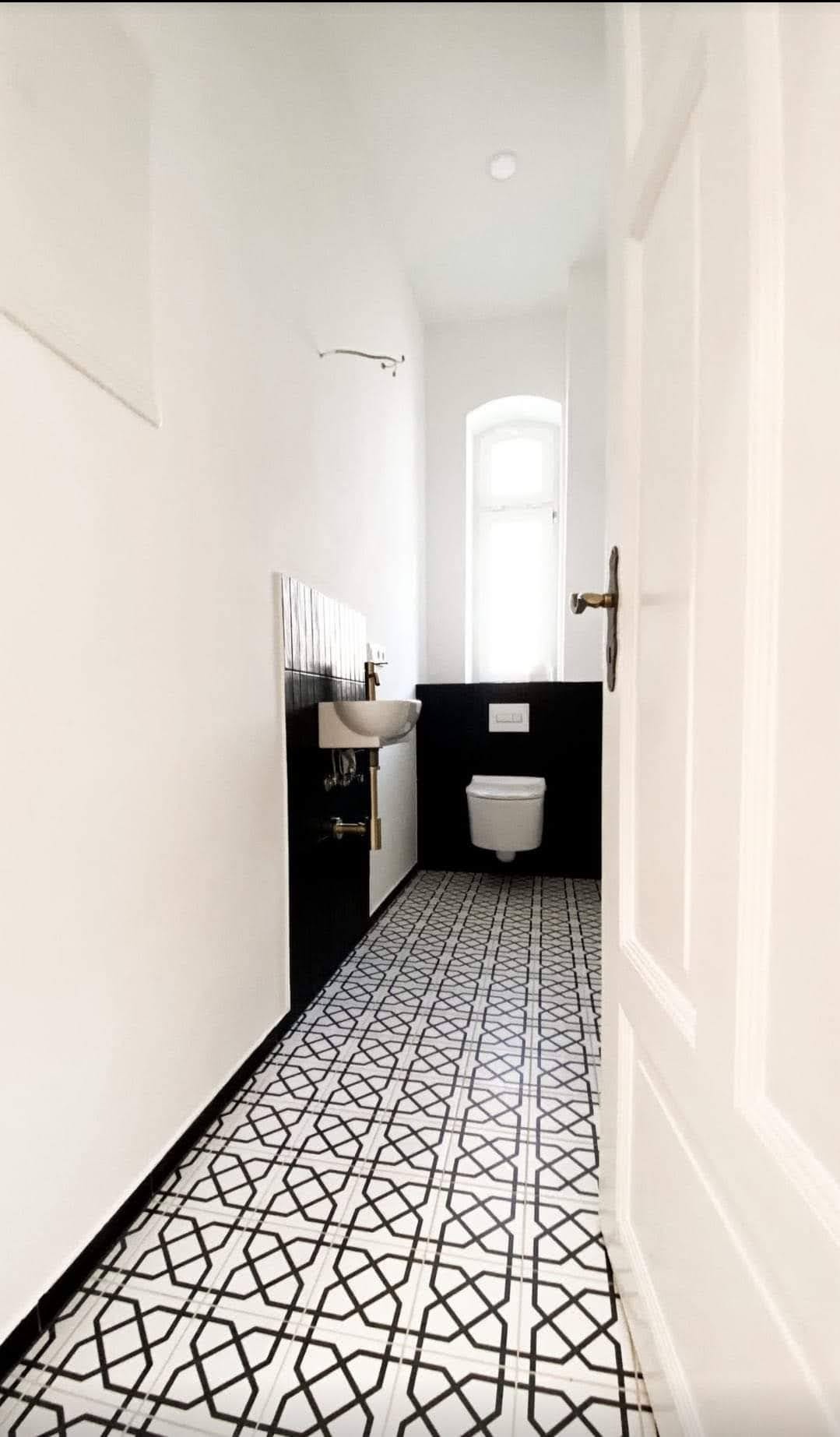 Wąska łazienka w bieli i czerni z geometryczną posadzką, umywalką nablatową i toaletą pod oknem, widok zza uchylonych drzwi.