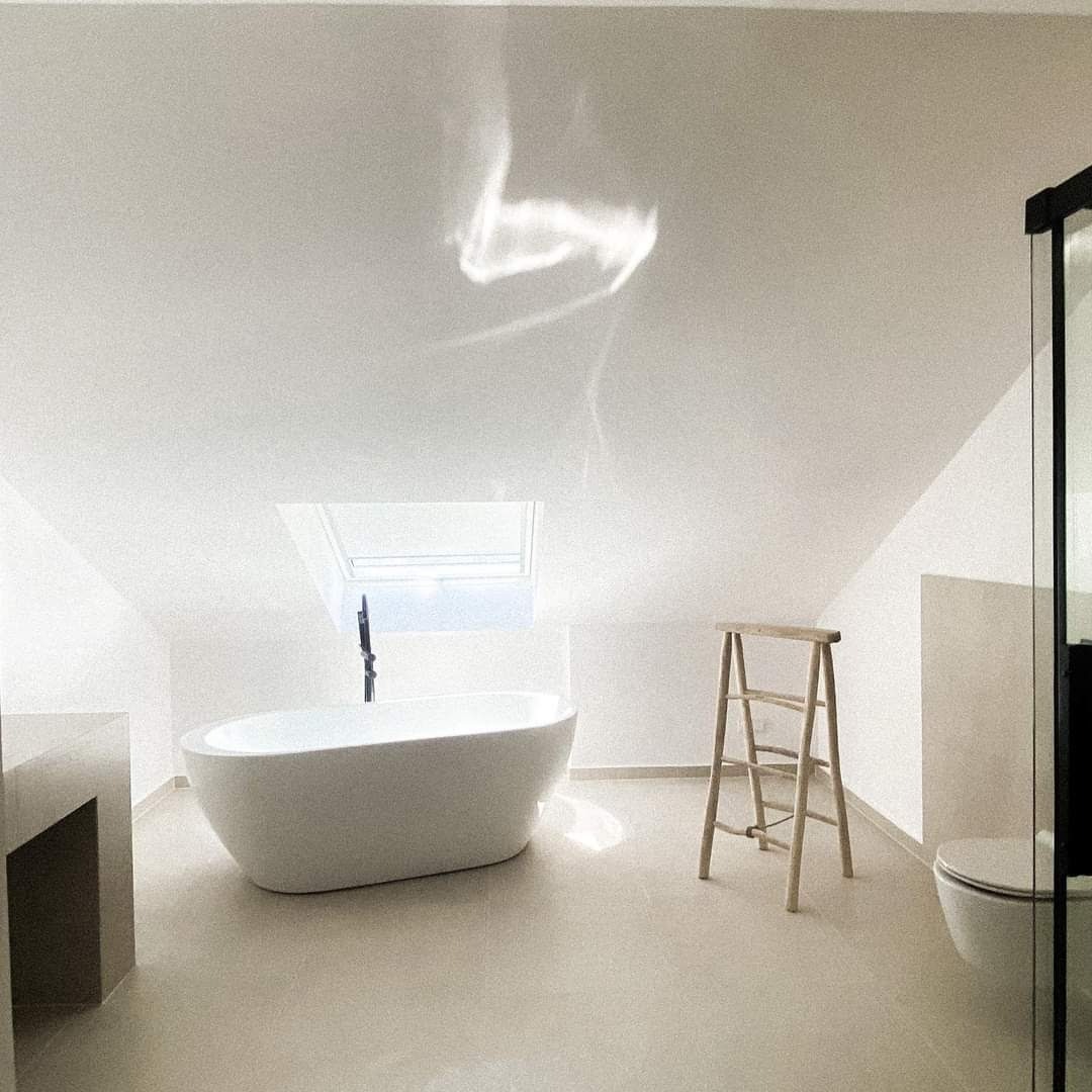 Jasna łazienka na poddaszu z wolnostojącą wanną, drewnianą drabinką i oknem dachowym, w minimalistycznym stylu, z refleksami światła na suficie.