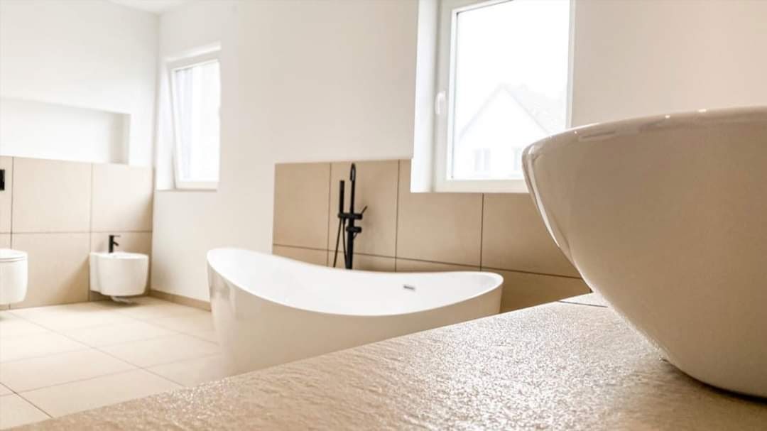 Nowoczesna łazienka z wolnostojącą białą wanną, czarną armaturą i beżowymi płytkami, umywalka nablatowa z bliska, widok na bidet i toaletę w tle, jasne wnętrze z oknami.