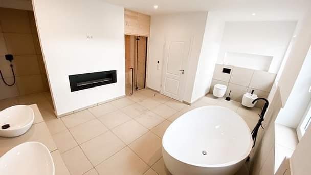 Przestronna łazienka w minimalistycznym stylu z wolnostojącą wanną, dwiema umywalkami nablatowymi, toaletą podwieszaną, grzejnikiem w ścianie i kabiną prysznicową ze szklanymi drzwiami.