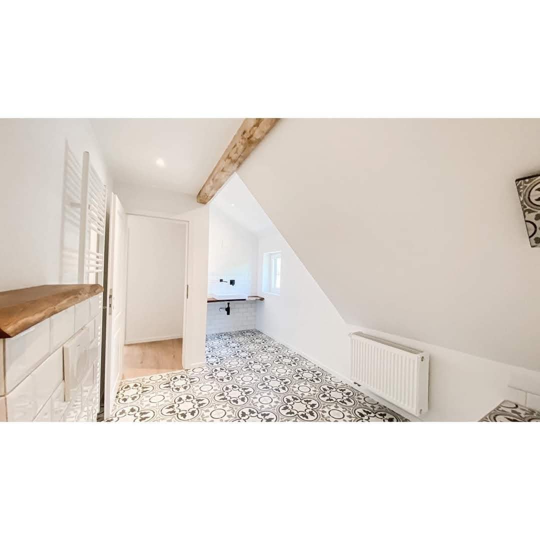 Białe wnętrze łazienki na poddaszu z dekoracyjną, wzorzystą podłogą, drewnianą belką stropową i grzejnikiem żeberkowym, widok na umywalkę na tle białych płytek.