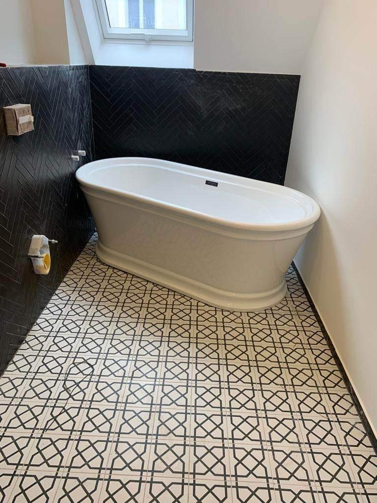 Wanna wolnostojąca w łazience z czarną, ułożoną w jodełkę glazurą na ścianach i wzorzystą, czarno-białą posadzką. Na ścianie widoczne pozostałości po kleju i gąbka.