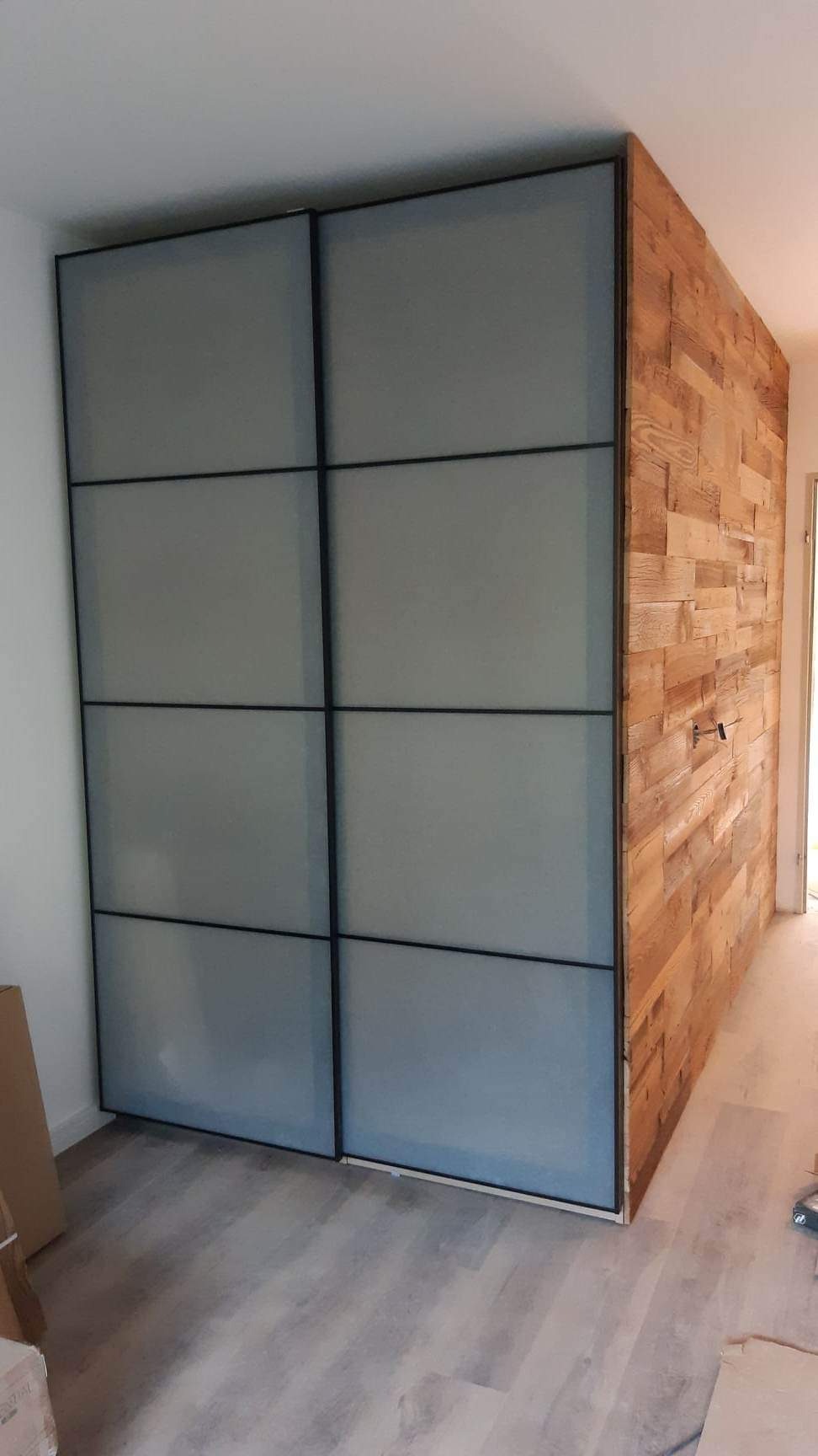 Szklana szafa przesuwna z czarnymi ramami, sąsiadująca z drewnianą ścianą z desek w pomieszczeniu z szarymi panelami podłogowymi.